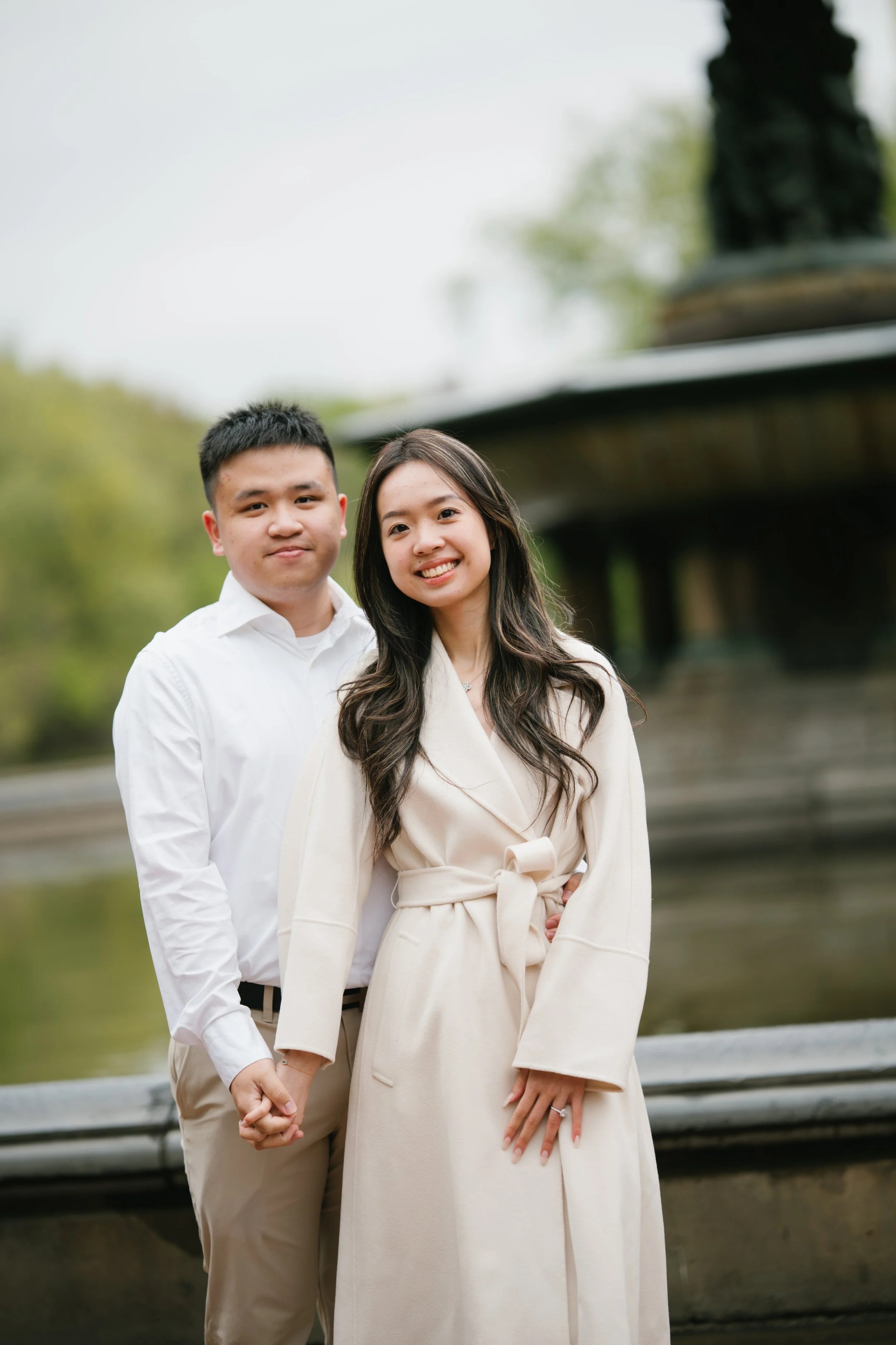 A young Asian couple stands outdoors holding hands, smiling, with trees and a metal fence in the background.