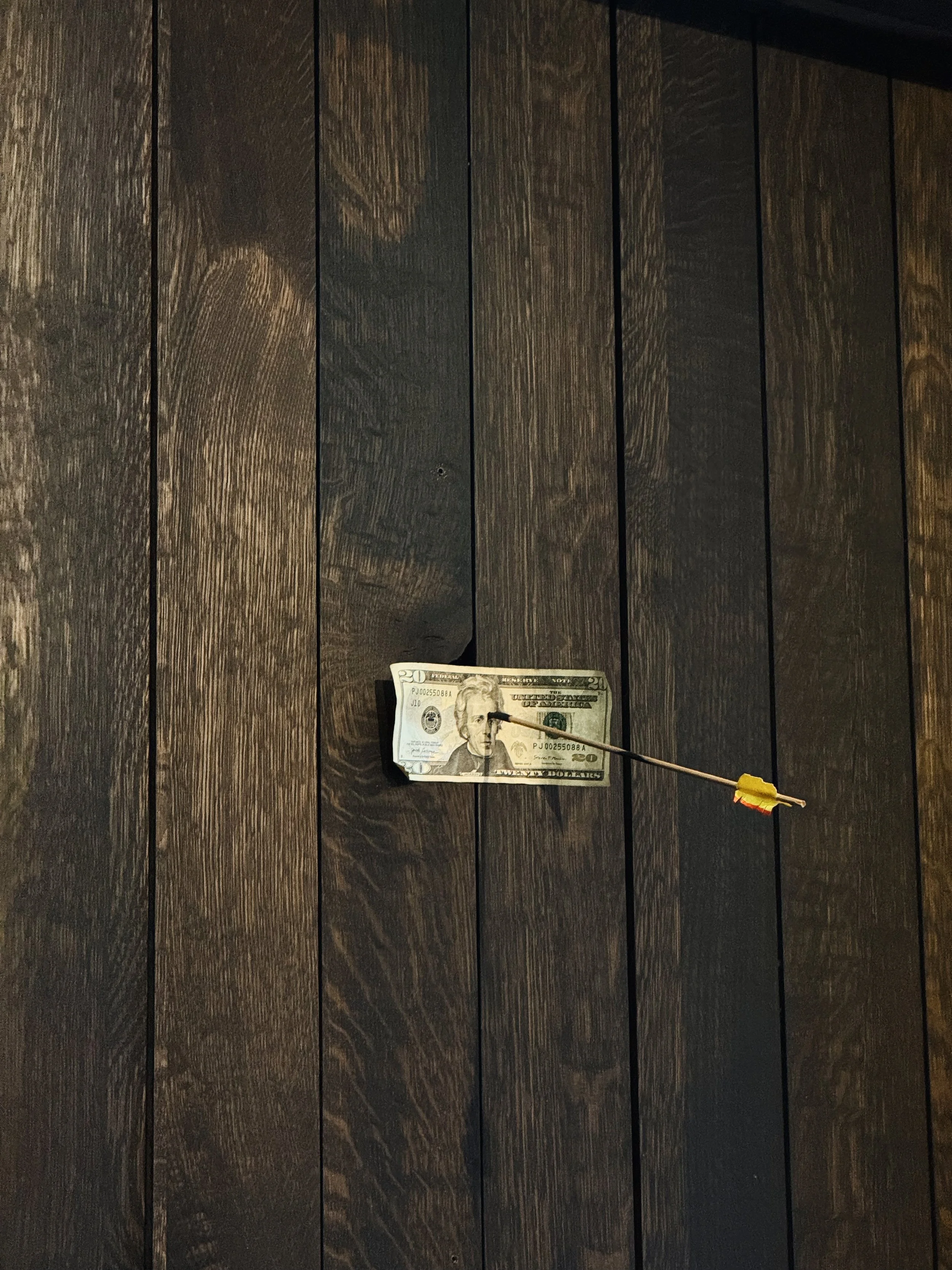 A twenty-dollar bill with a needle and a yellow clip attached to it, resting on a dark wood surface.