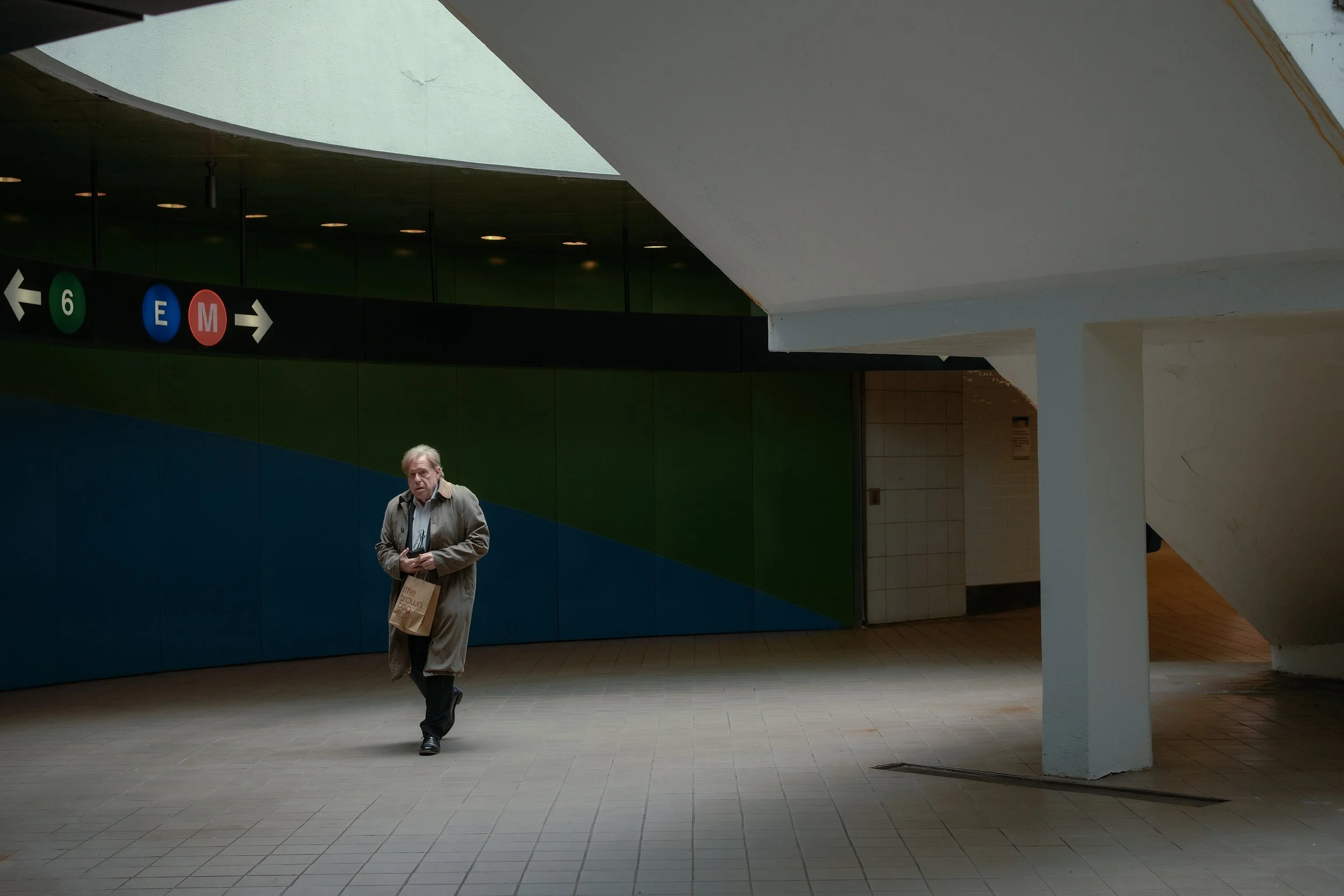 An older man walking through a modern, indoor space with colorful walls and ceiling, holding a paper bag and a small object in his hands.