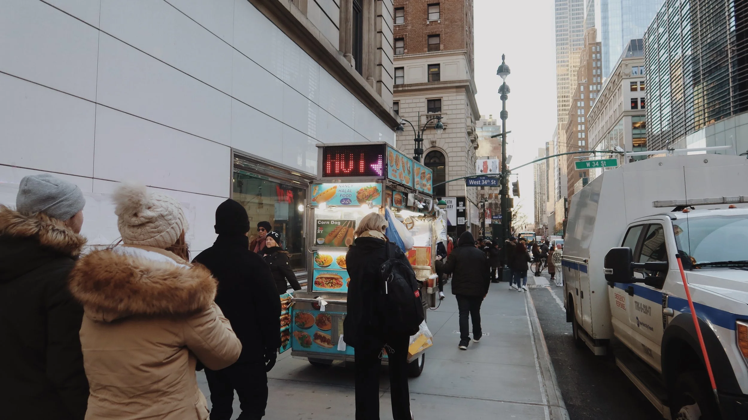 Crowded city sidewalk with people wearing winter coats and hats, standing near a hot dog stand. In the background are tall skyscrapers and a street sign for West 34th Street.