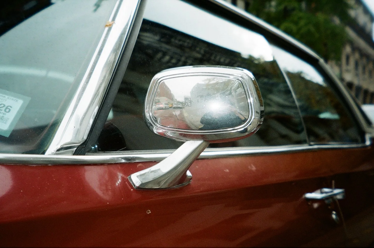 Close-up of a classic red car's side mirror and part of the windshield, with a reflection of a street scene and sky.