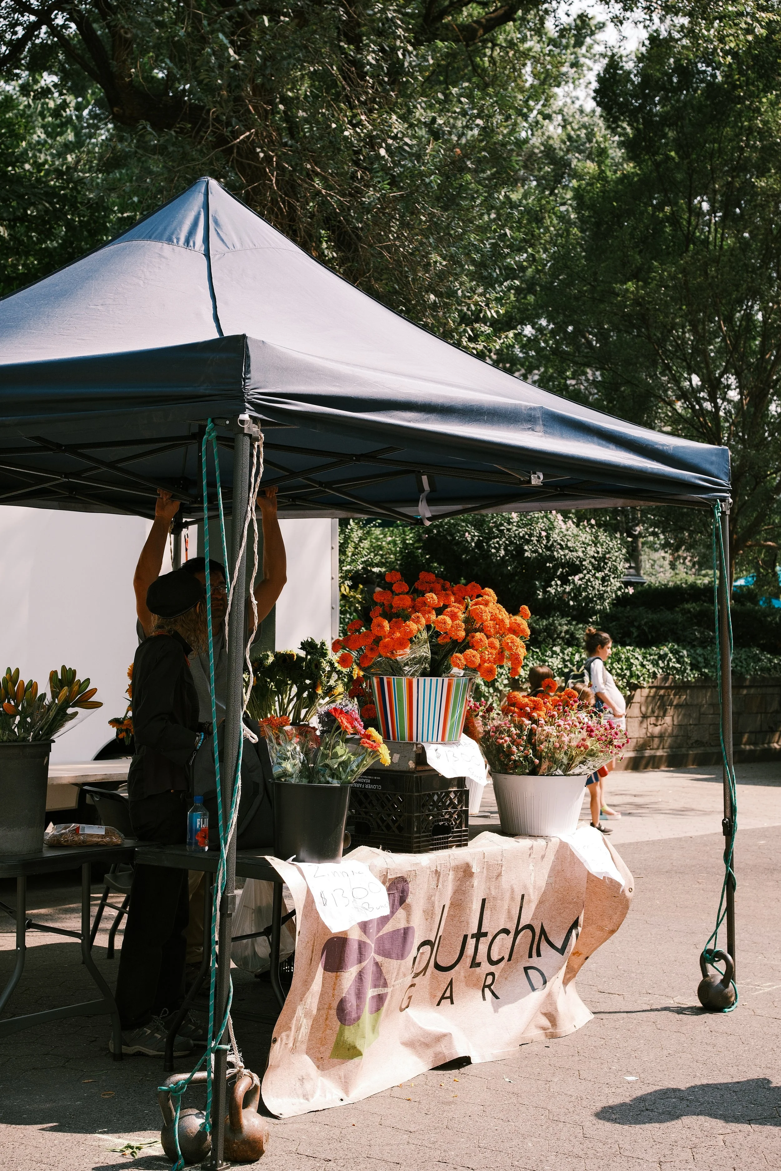 A flower stand at an outdoor market with various colorful flowers, including orange marigolds, displayed in baskets and pots. A person is visible behind the stand, holding onto the tent pole, with trees and pedestrians in the background.