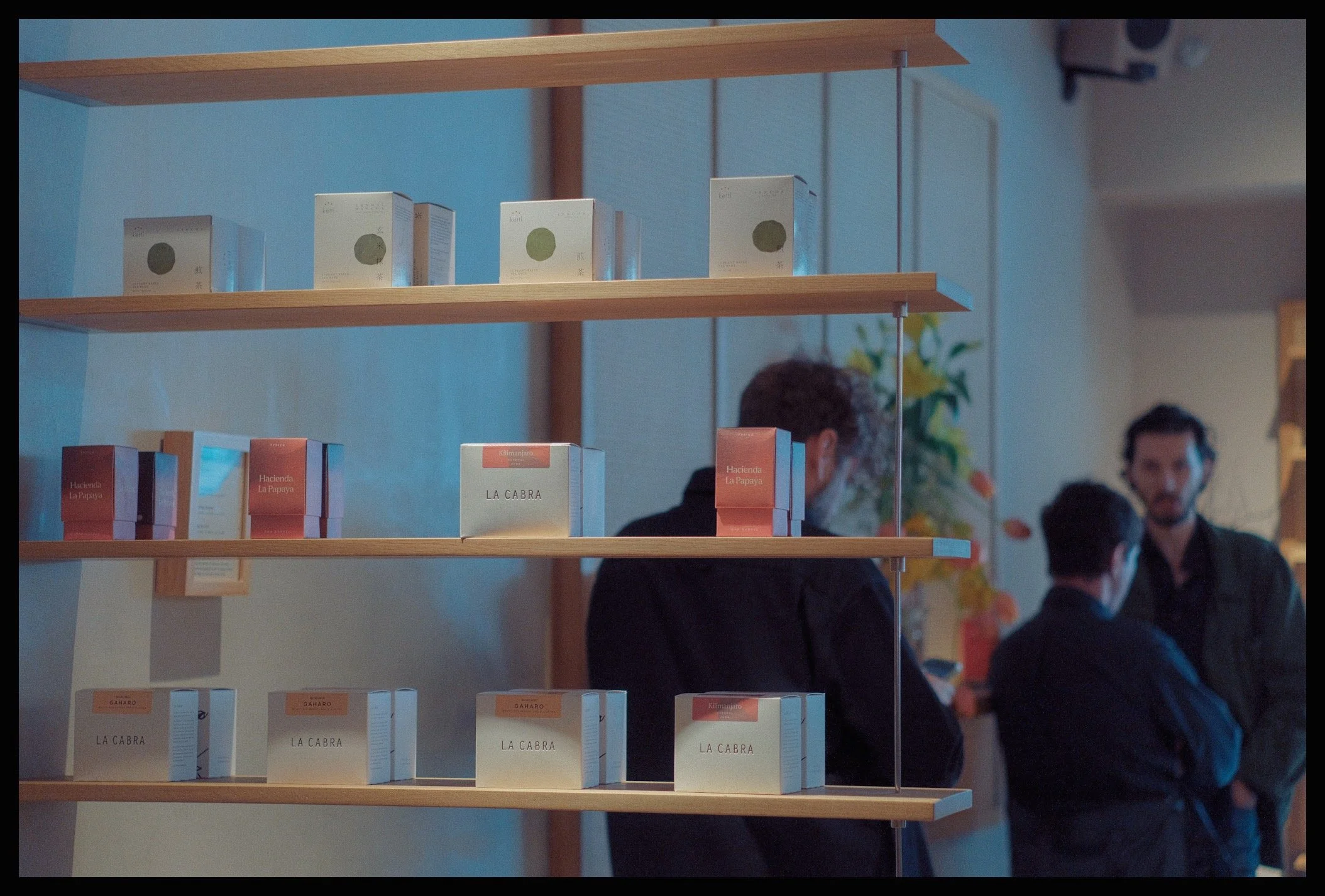 Wooden shelves with small boxes labeled "La Cabra" and other text, set in an indoor space with people in the background.
