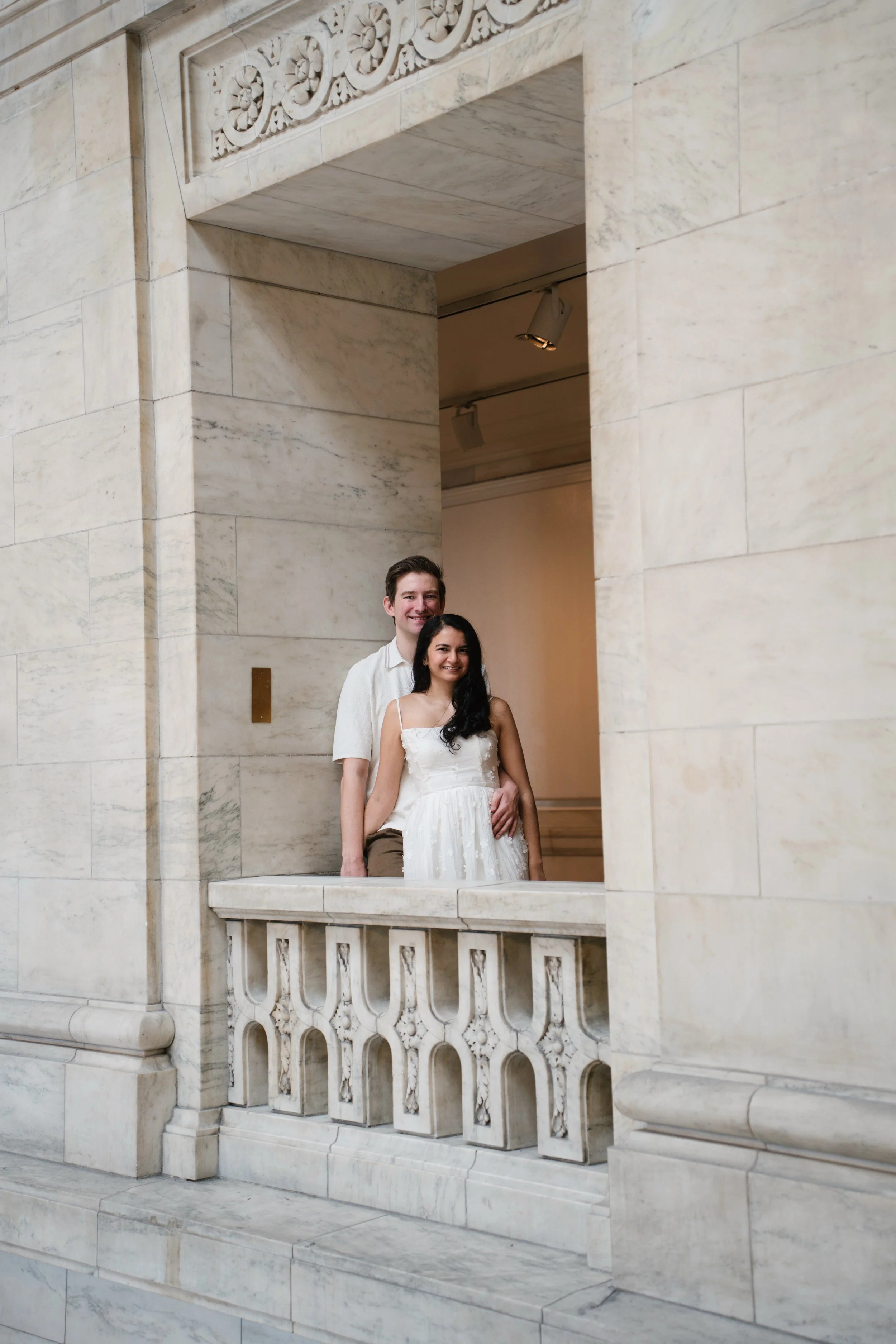 A couple dressed in wedding attire standing on a marble balcony of a historic building with ornate architectural details.