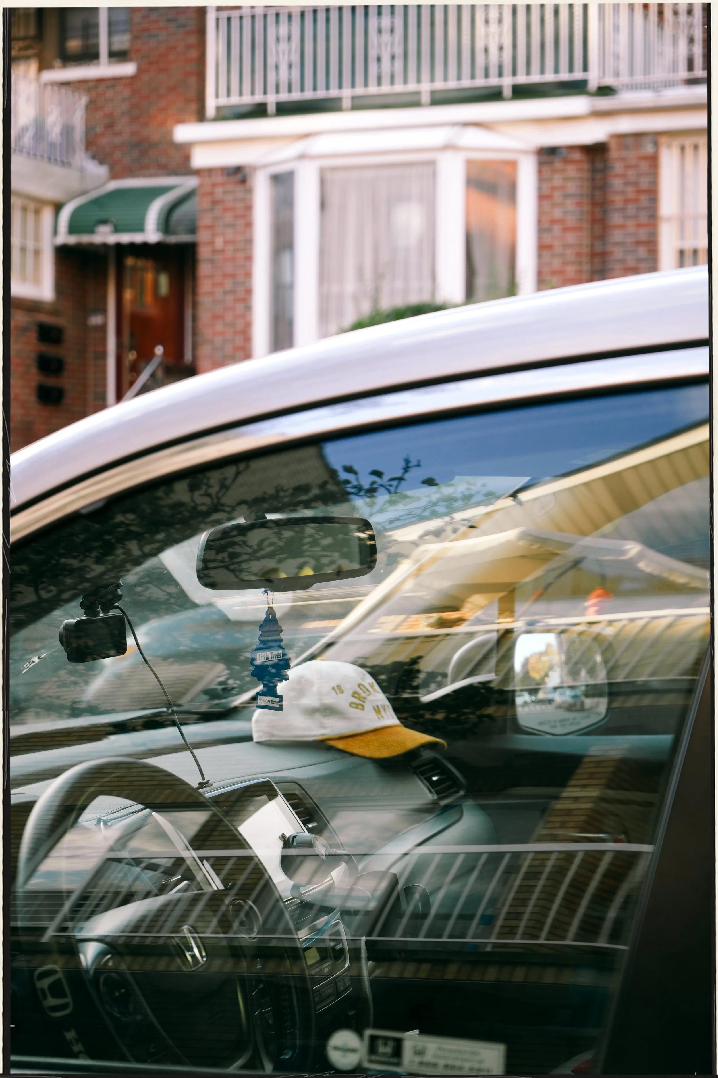 The image shows the interior of a car with a view through the windshield. There are multiple reflections and overlapping layers, including parts of other parked cars and a building with brick walls and balconies in the background. A hanging air fresh