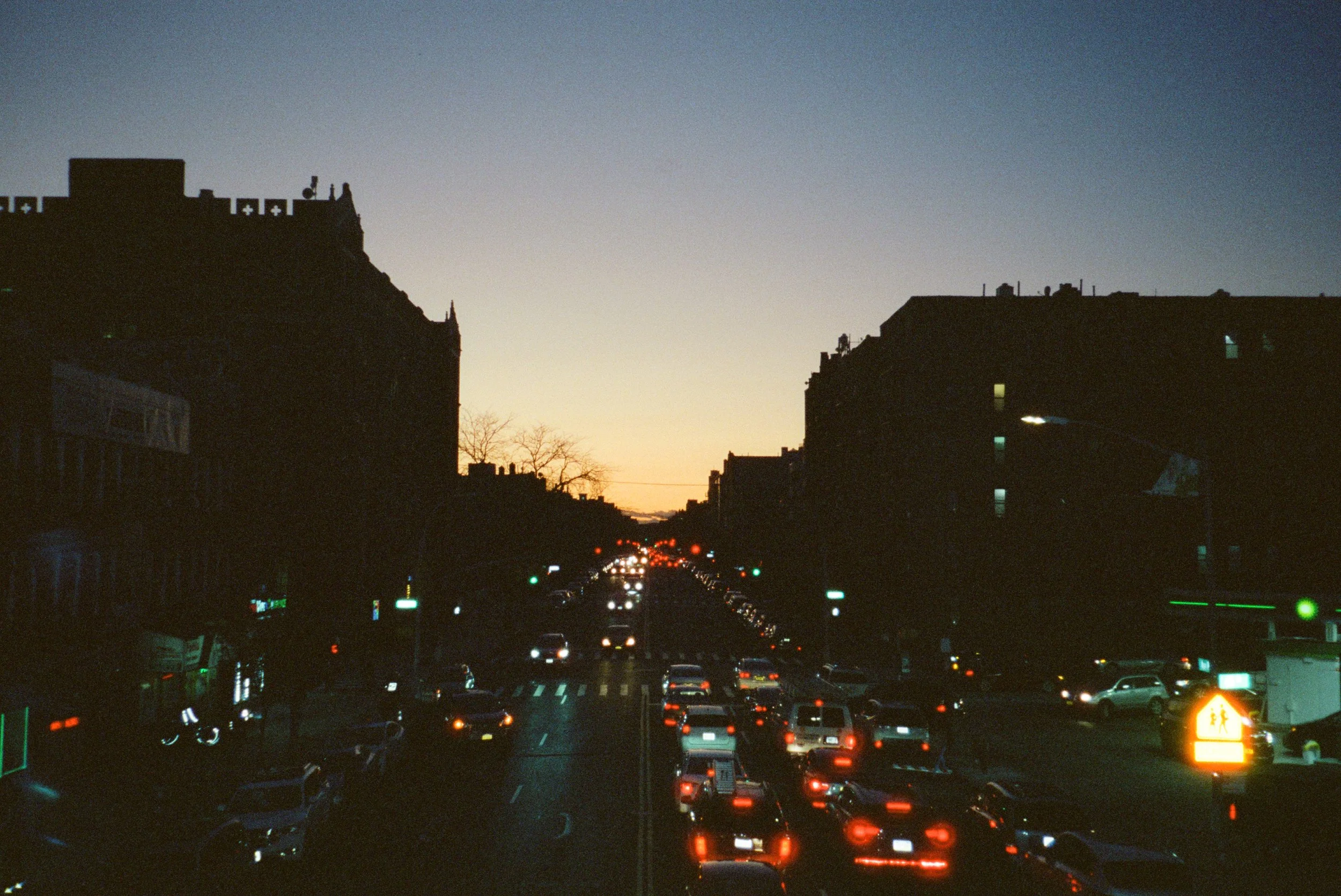City street filled with moving cars at dusk, silhouetted buildings on either side, and a cloudy sky with a faint sunset glow.