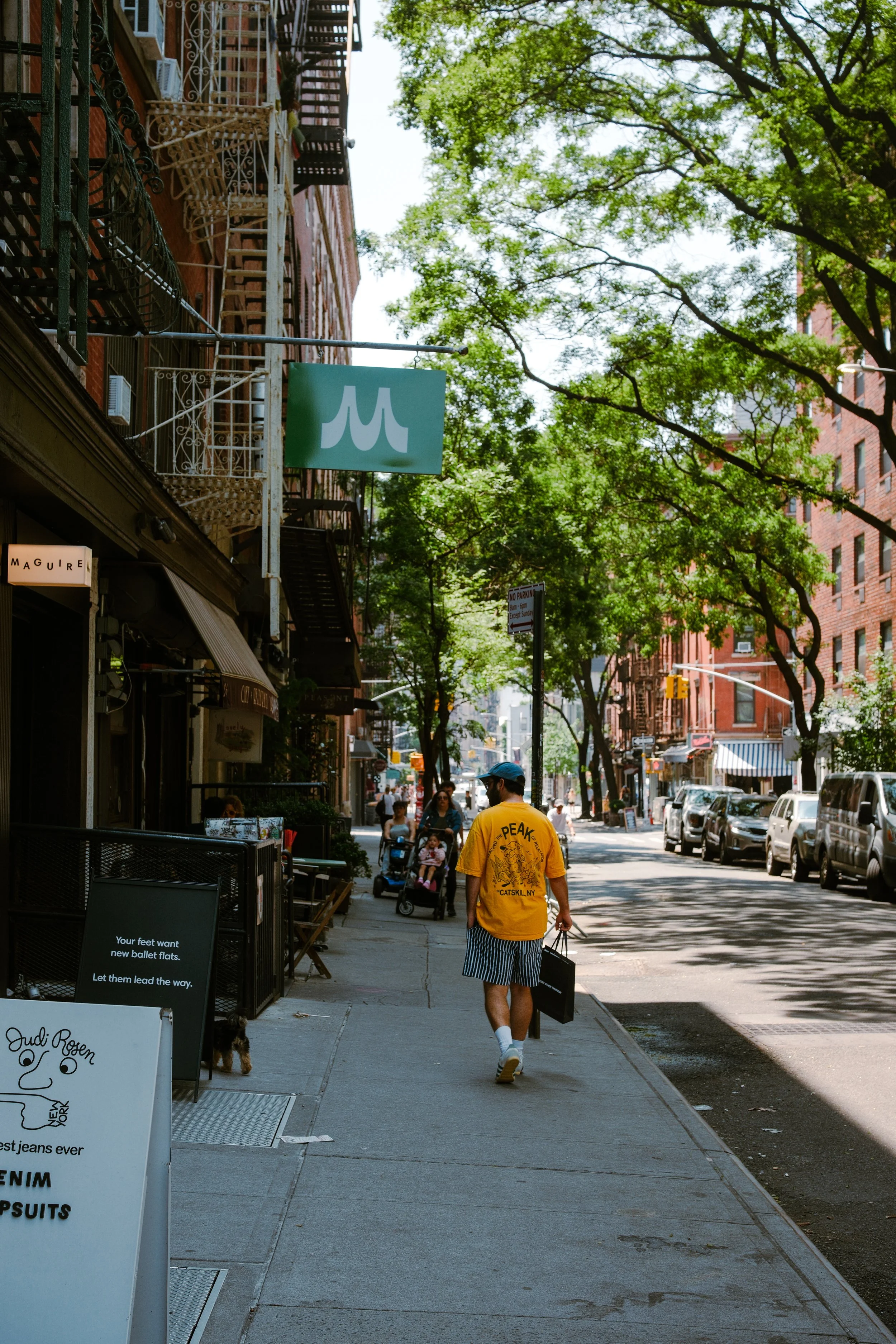 A city sidewalk lined with trees and brick buildings. People are walking, including a man in a yellow shirt with a tattoo and striped shorts, carrying shopping bags. There are parked cars on the street, and storefront signs visible, including one wit
