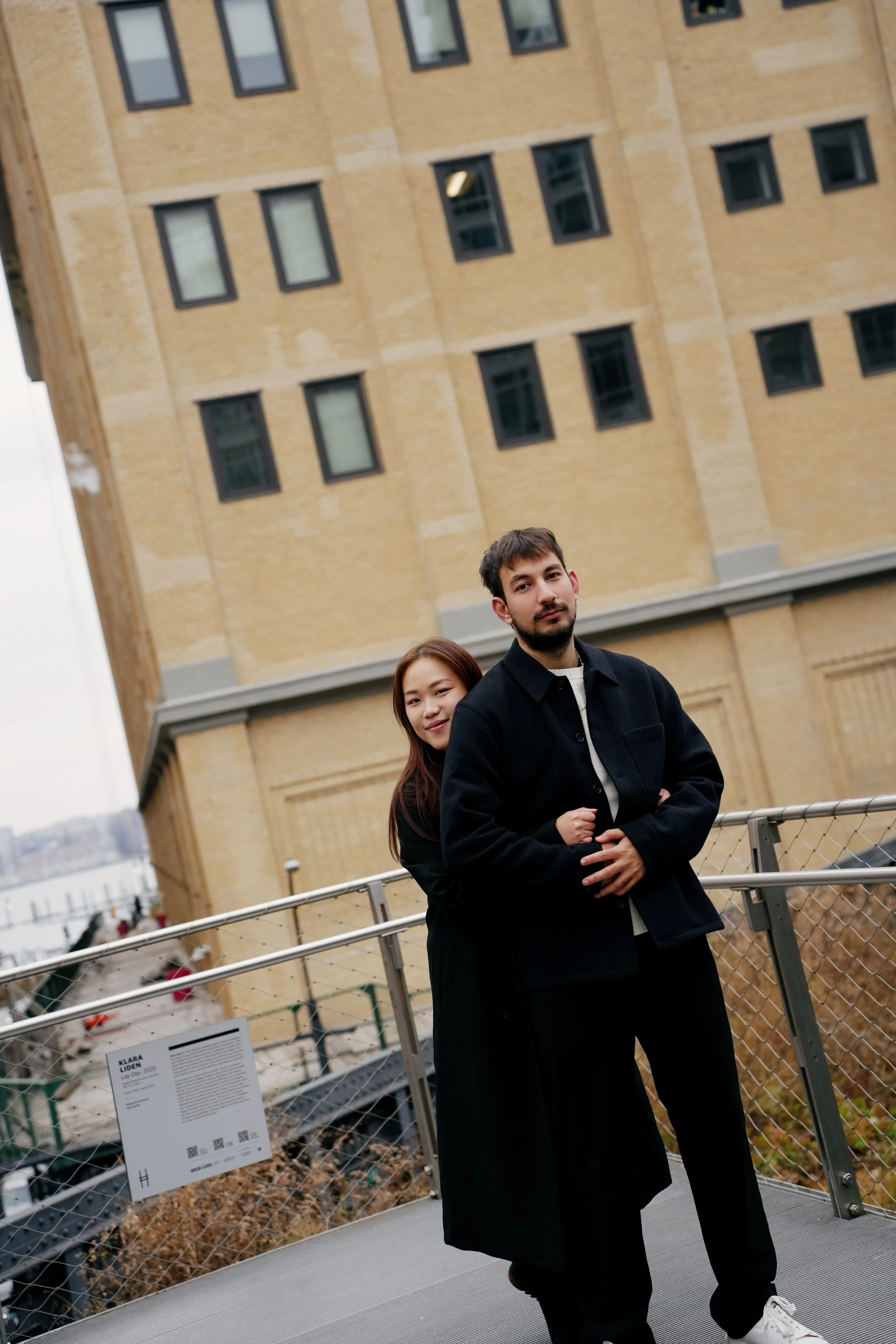 A man and a woman standing on a bridge over a canal with boats, in front of a large yellow brick building with multiple windows.