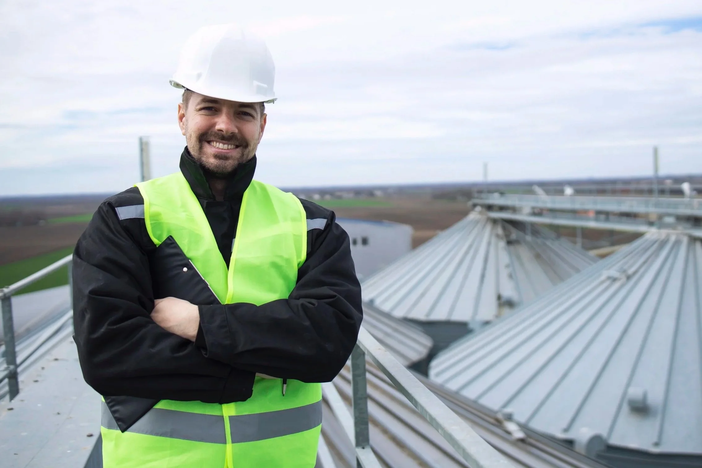 A man wearing a white safety helmet and a reflective yellow safety vest stands outdoors with arms crossed, smiling. Behind him, there are metal mechanical structures and a cloudy sky.
