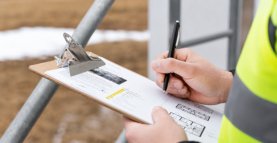 Person in a yellow safety vest writing on a clipboard with a pen at a construction site.