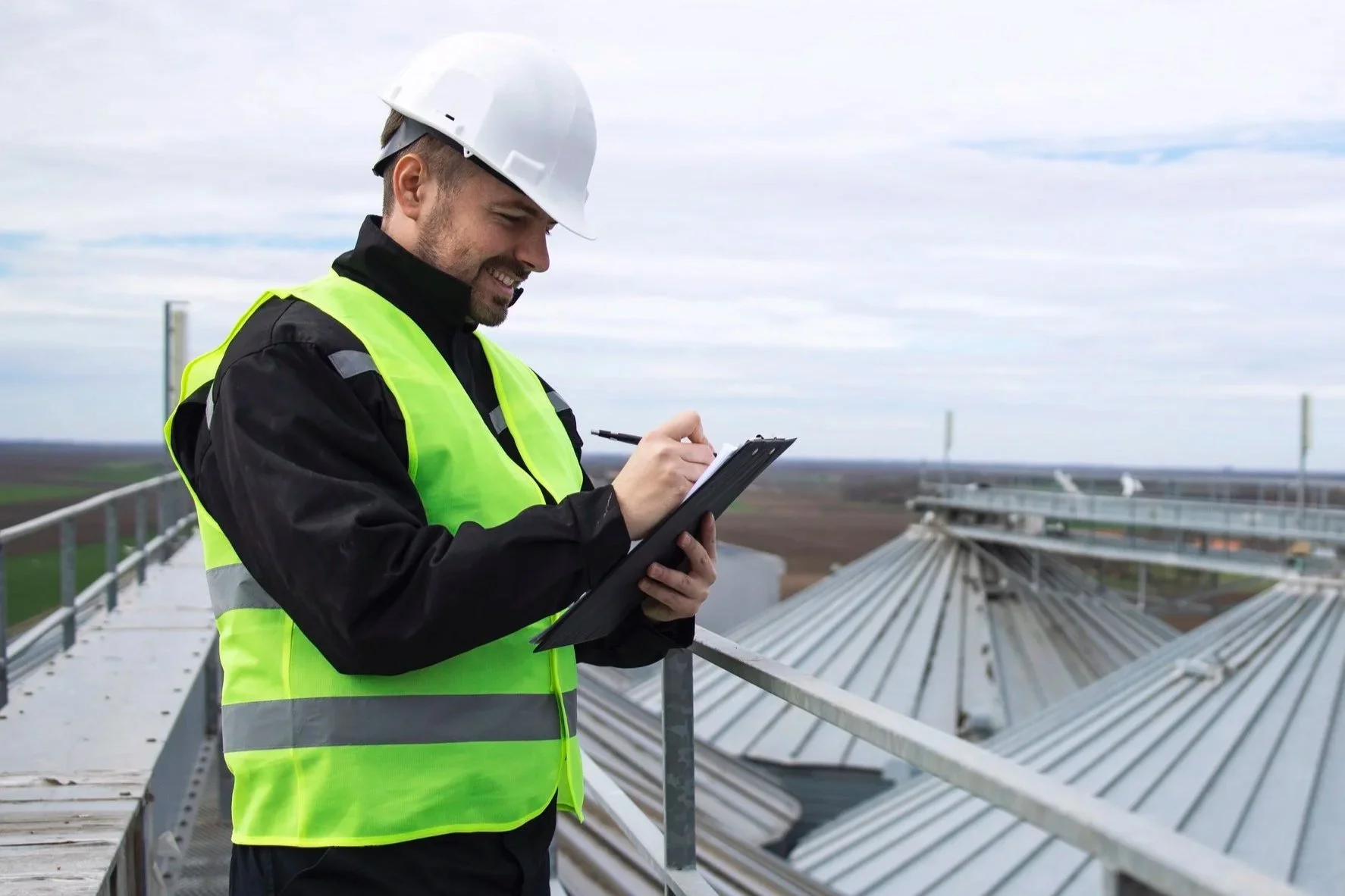 A man wearing a yellow safety vest and white hard hat writing on a clipboard on a roof or industrial facility outdoors.