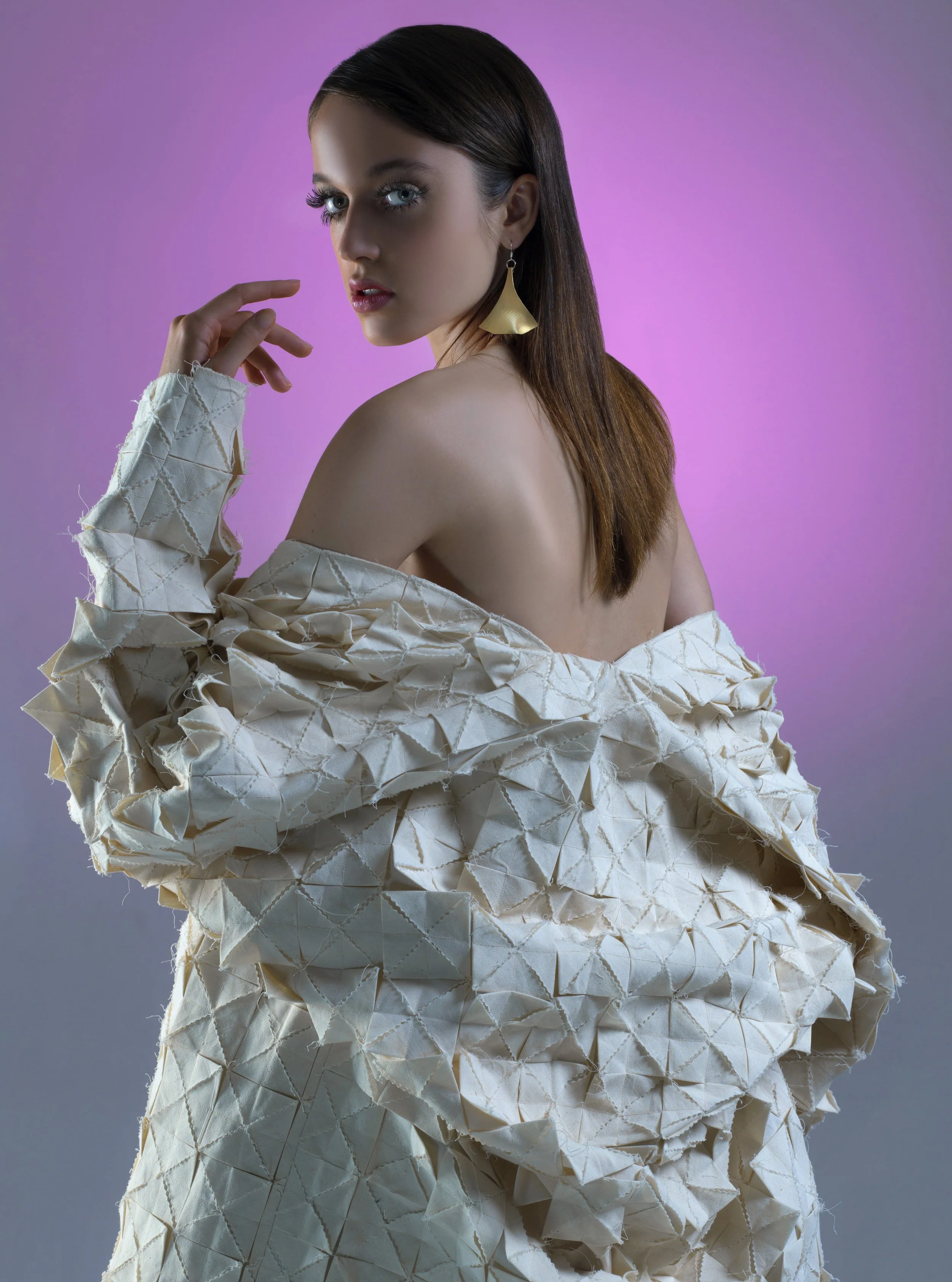 A woman with long brown hair and gray eyes, wearing large gold earrings and a textured off-shoulder cream jacket, posed against a purple gradient background.