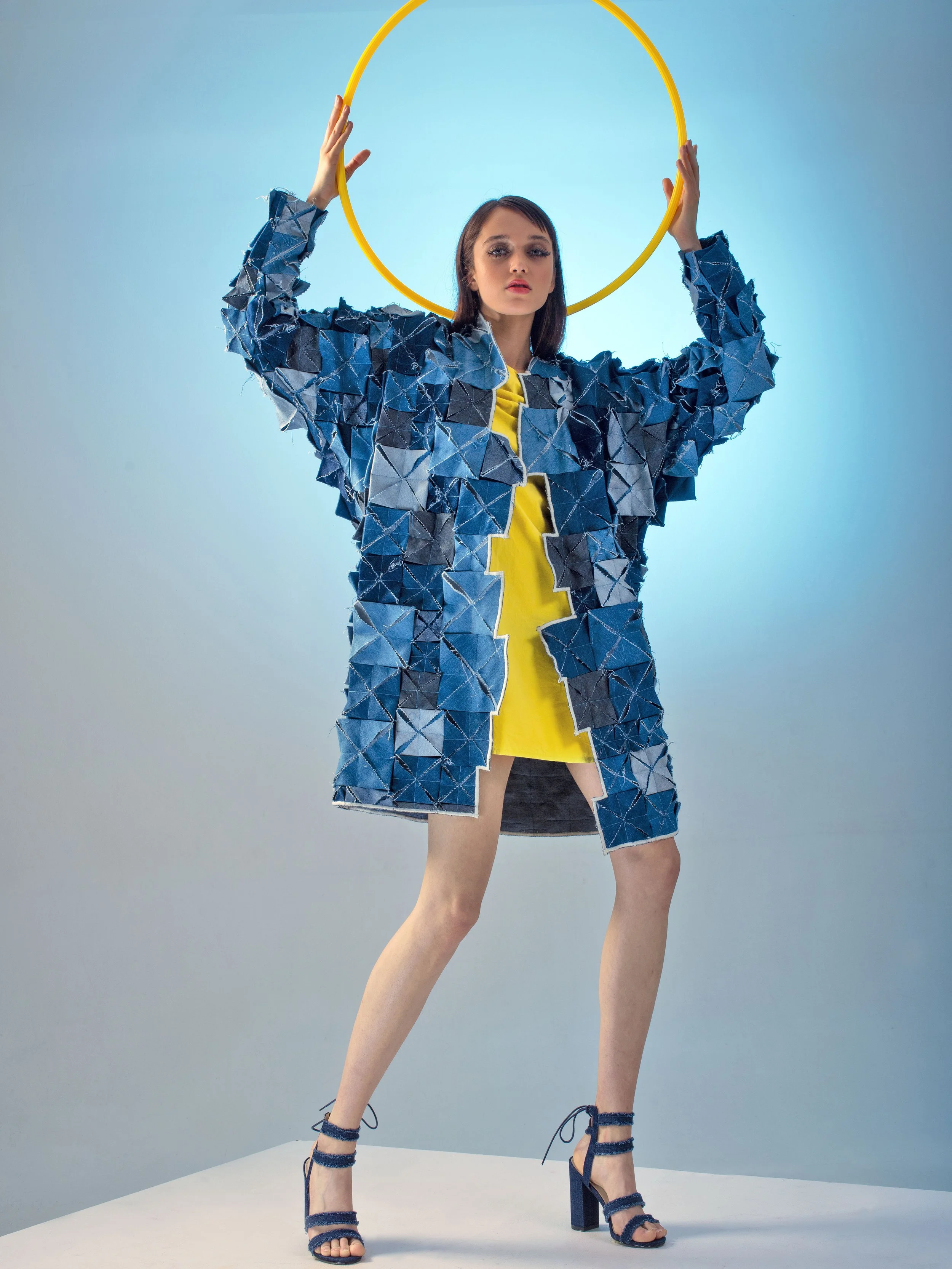 A woman standing with one leg forward, wearing high-heeled blue shoes, a yellow dress, and a patchwork blue denim coat, holding a yellow hula hoop above her head against a light blue background.