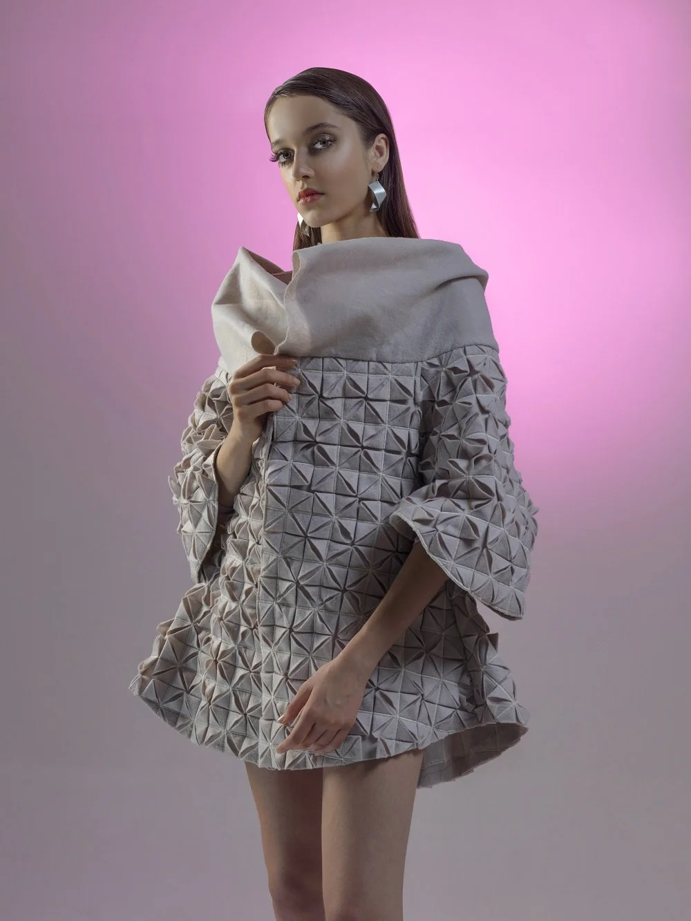 A woman with long brown hair wearing a beige, textured dress with geometric patterns and a wide collar, posing against a pink and purple gradient background.