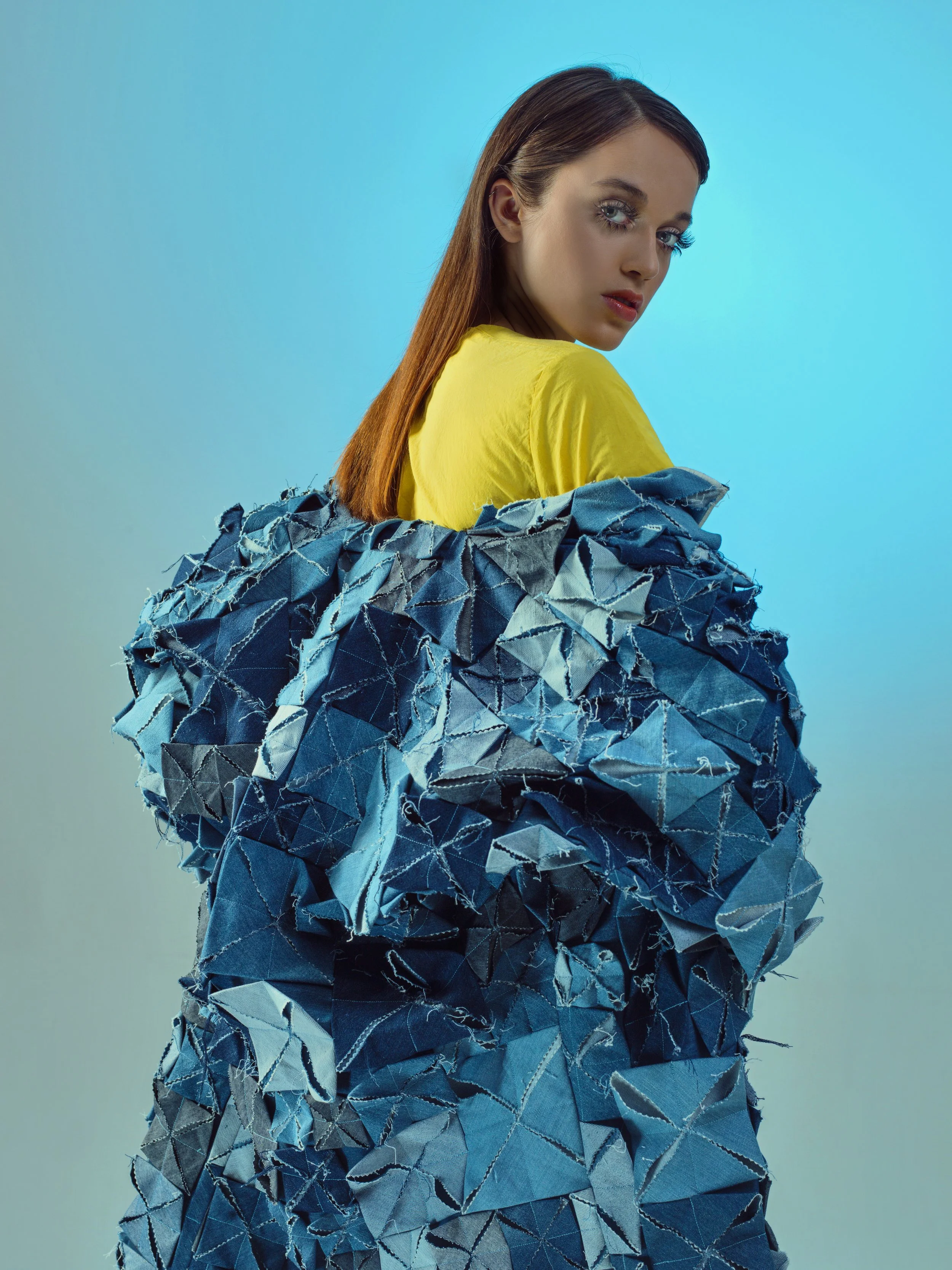 A woman with long brown hair, wearing a yellow top, is wrapped in an oversized, textured blue jacket made of torn fabric pieces against a light blue background.