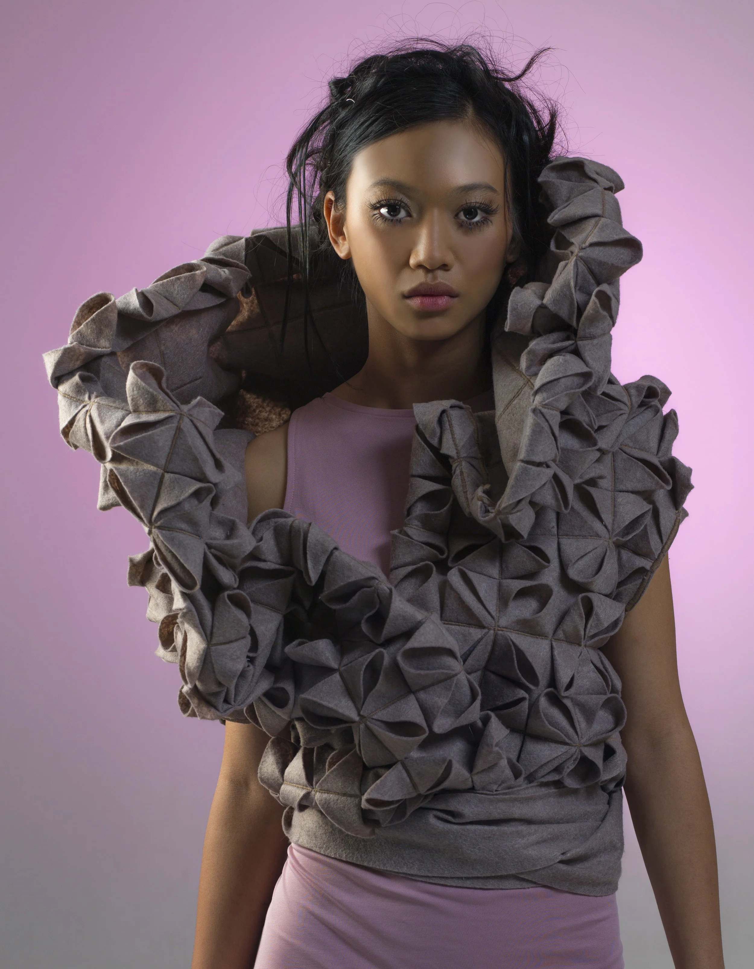 A woman with dark, messy hair, wearing a gray textured jacket over a light purple top, is posing against a pink and purple background. She has a serious expression and is looking directly at the camera.