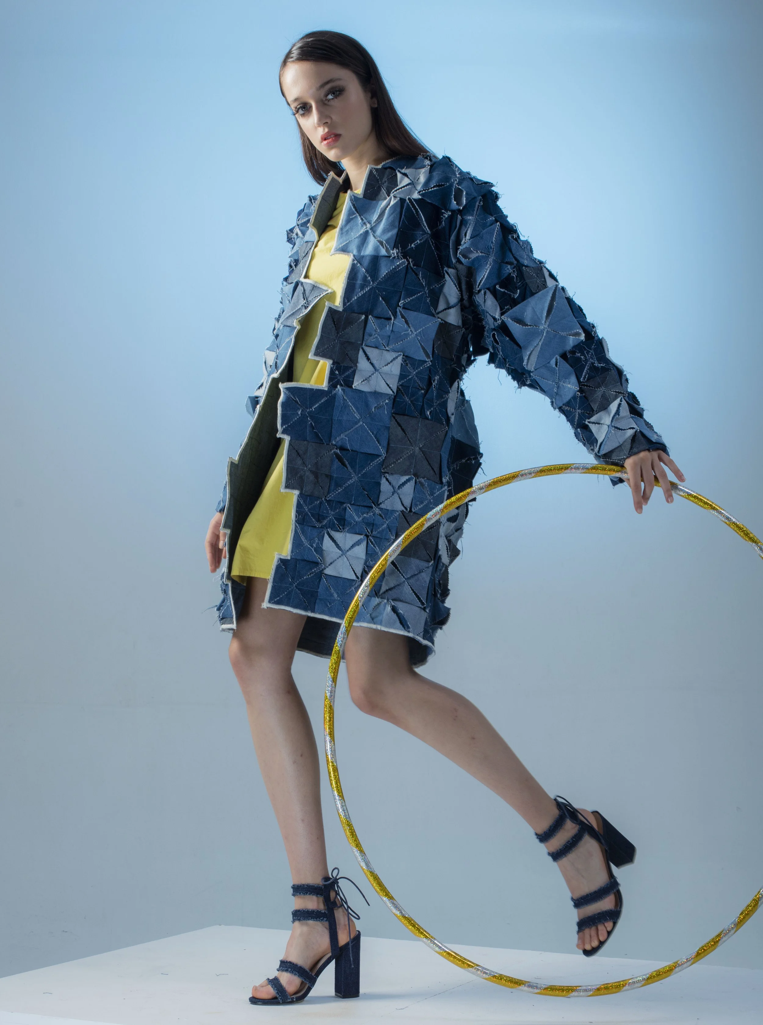 Fashion model wearing a textured blue coat with a yellow dress underneath, high heels, standing on one leg, holding a gold and silver hula hoop, against a plain light blue background.