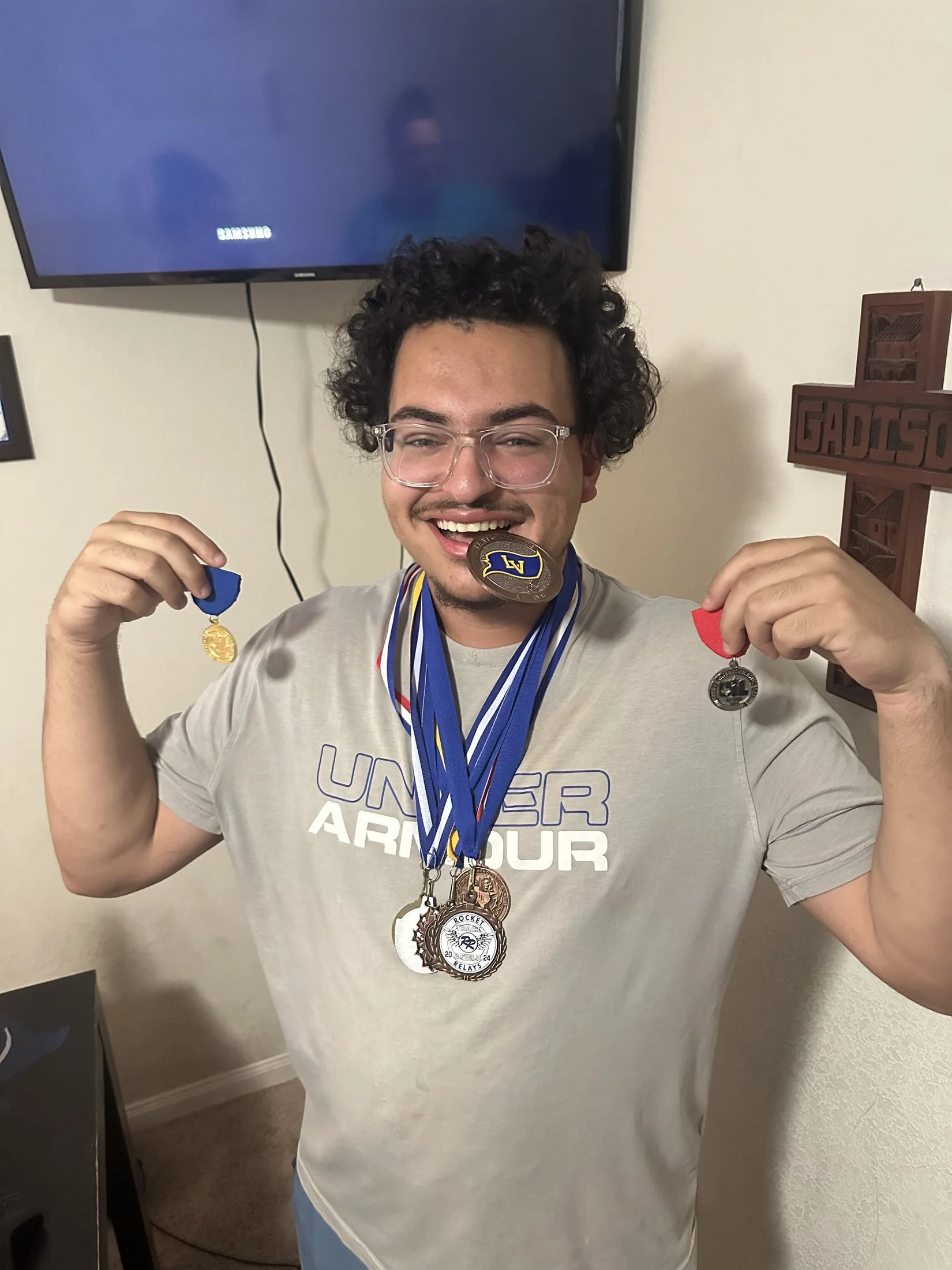 A young man with curly dark hair and glasses smiling while holding up medals around his neck. He is wearing a gray T-shirt with the word 'UNDER ARMOUR' and a logo on it. There is a television on the wall behind him displaying the Samsung logo and a w