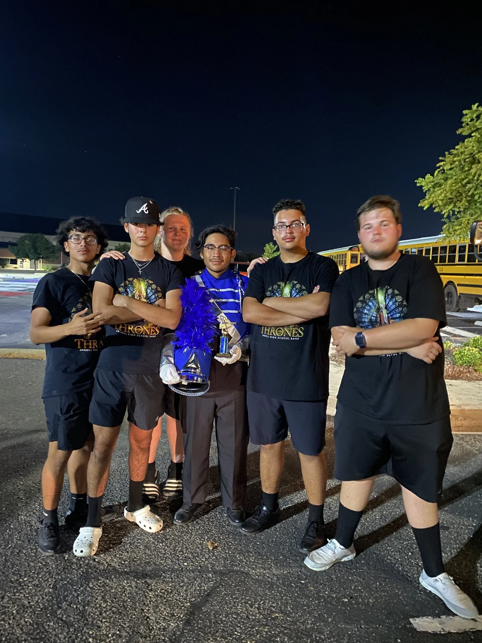 Group of six young men standing outdoors at night, some with arms crossed, one holding a trophy, in front of a yellow school bus and trees, with a dark sky overhead.