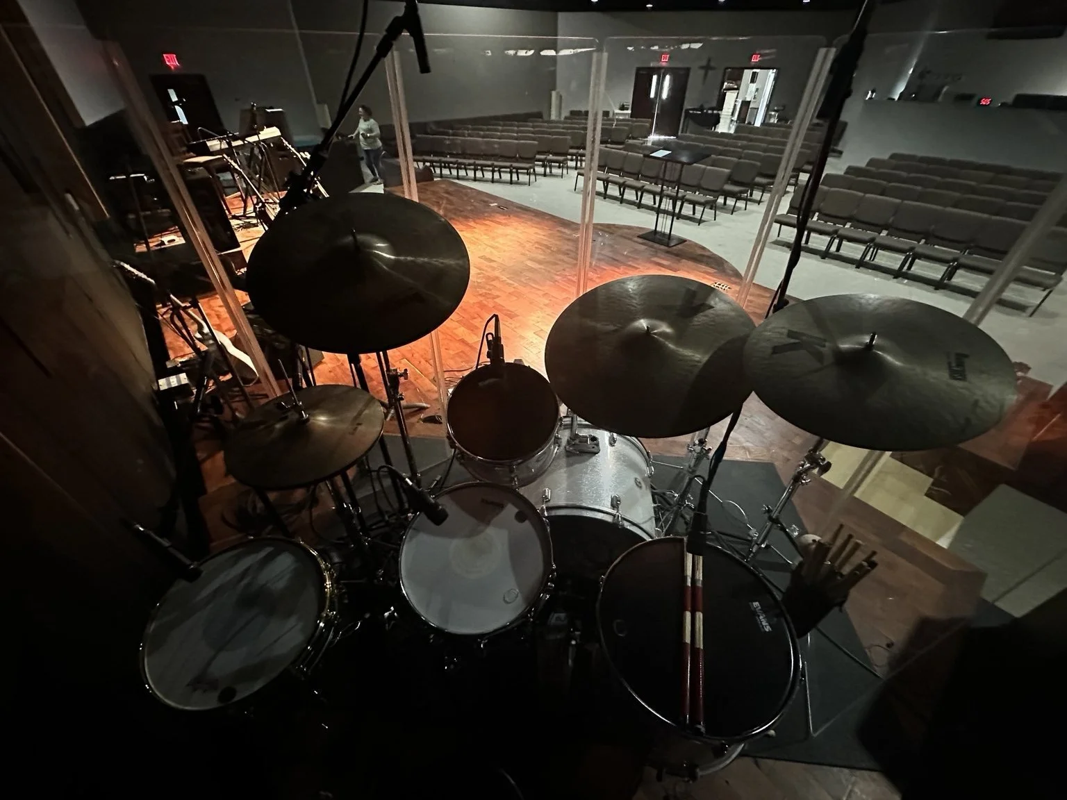 View from behind a drum set on stage, overlooking an empty auditorium with rows of seats, microphones, and a person in the background.