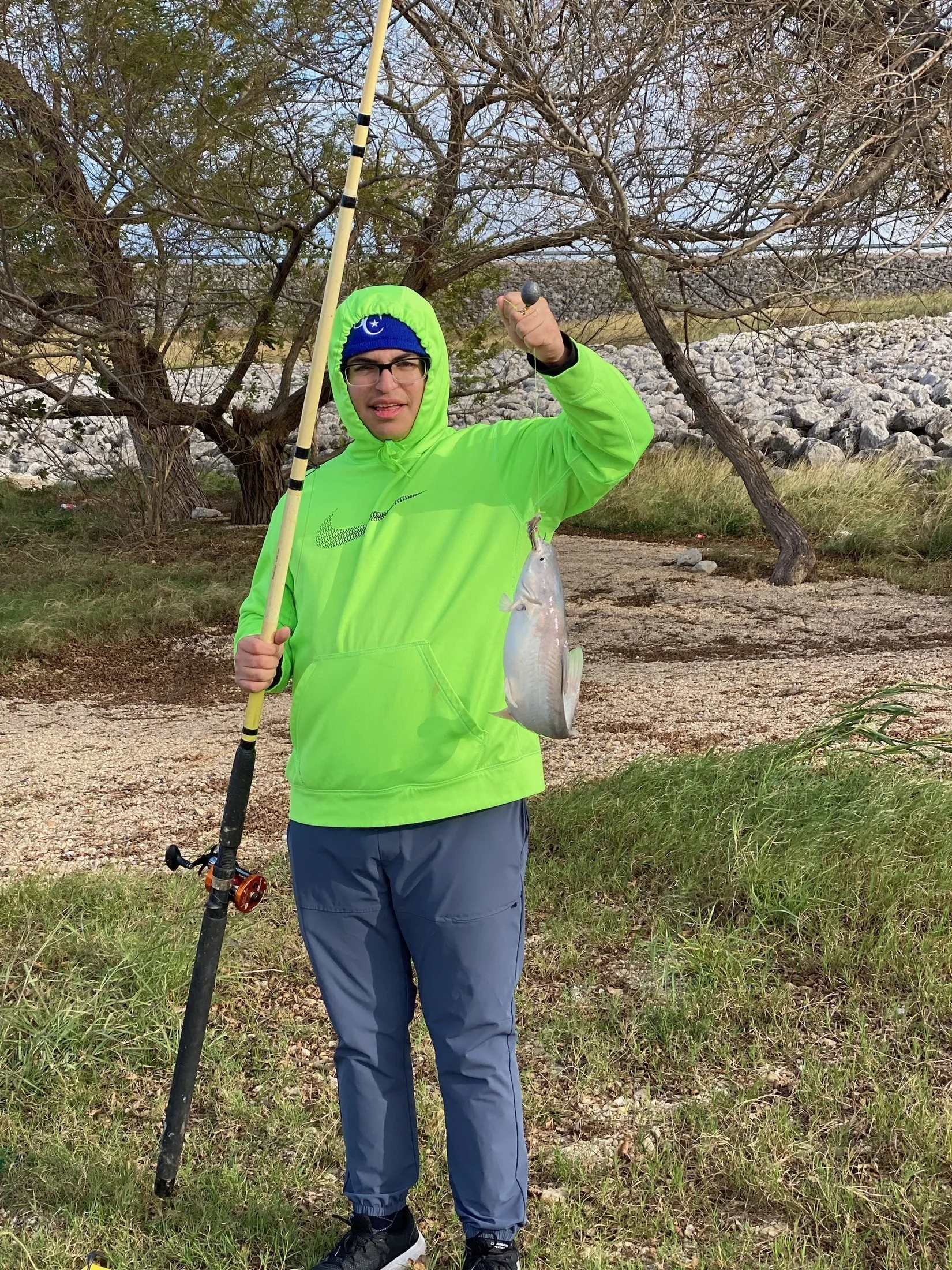 Man wearing a bright green hoodie and glasses holding a fishing rod in one hand and a fish in the other, standing outdoors on a grassy area with a tree and rocks in the background.