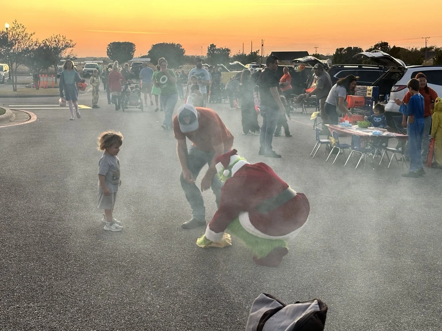 People gathered outdoors during sunset, with a child interacting with a person dressed as the Grinch and another dressed as Santa Claus, in front of various tables and vehicles, during a holiday event.
