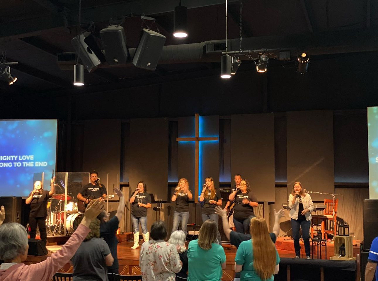 A church service or worship event with a band and choir singing on stage, congregation members raising hands, and a large illuminated cross on the back wall.