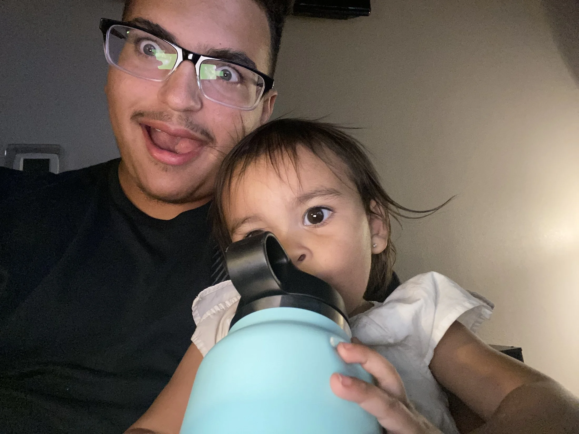 A man with glasses and a mustache showing a surprised or playful expression, holding a teal-colored thermos, with part of a young girl's face and shoulder visible in front of him.