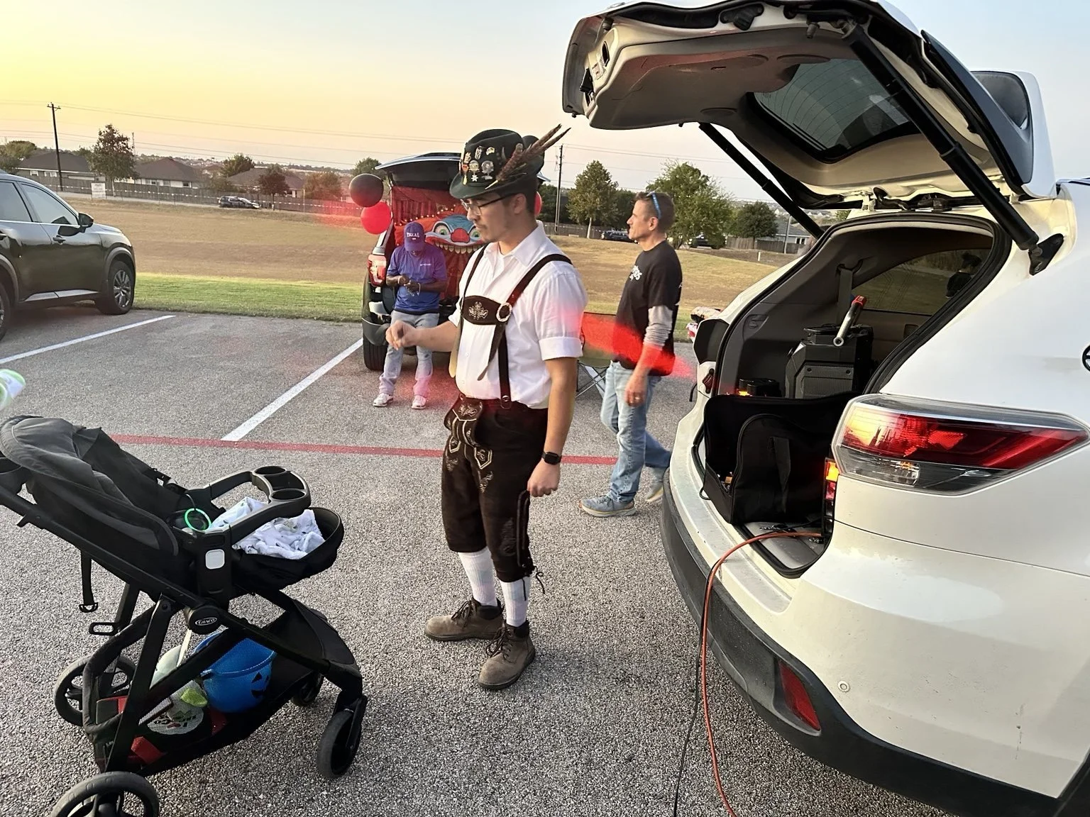 A man dressed in traditional Bavarian clothing with a feathered hat, standing next to a white SUV with its trunk open, at an outdoor event in a parking lot during sunset. A stroller with a baby inside and other people are also visible in the backgrou