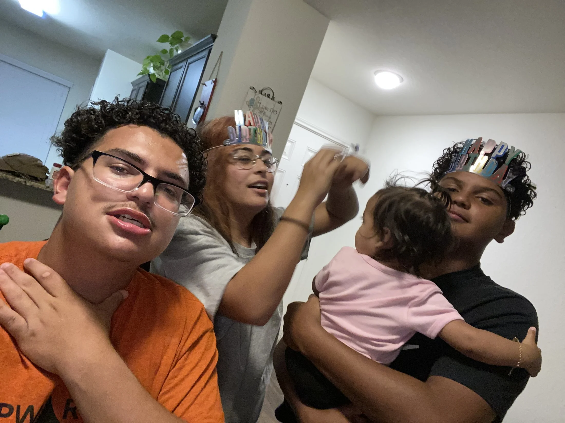 Group of five people celebrating a birthday, wearing colorful 'Happy Birthday' headbands, in a home setting with white walls and ceiling lights.