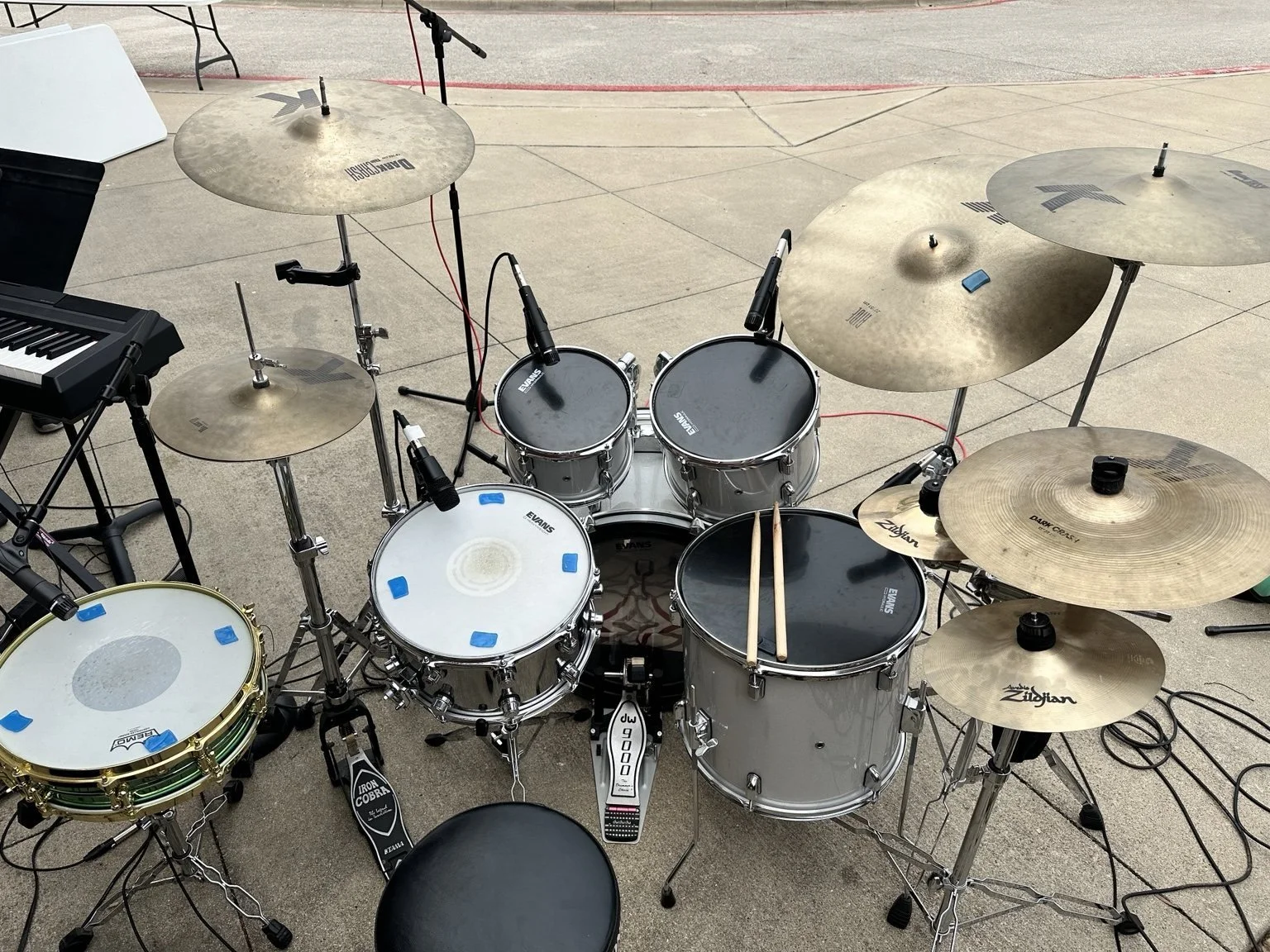 A drum set with various cymbals and drums arranged on a concrete stage, accompanied by a keyboard on a stand, with microphone stands and cables nearby.
