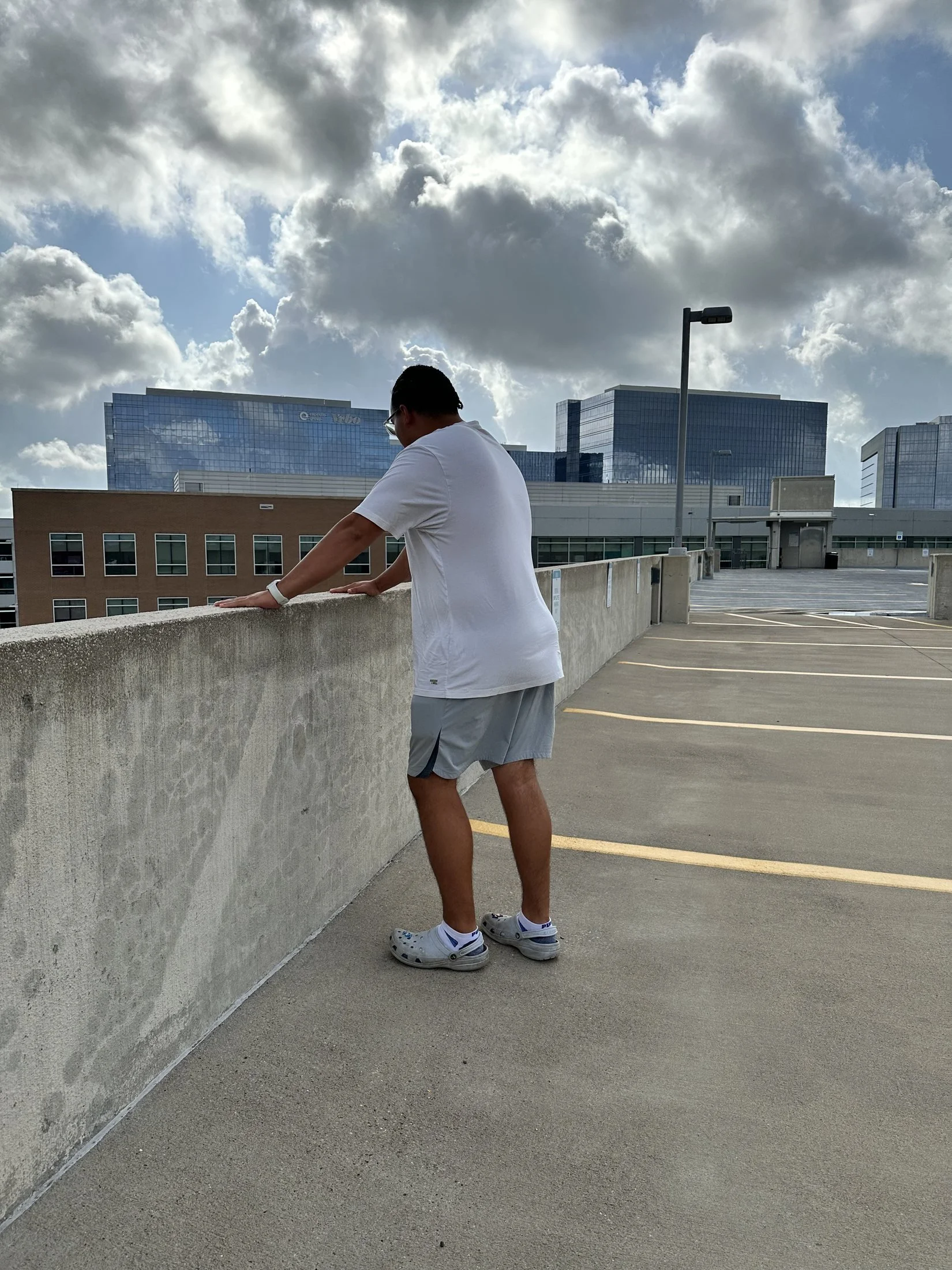 A man leaning on a concrete barrier on a rooftop parking lot with modern glass buildings and cloudy sky in the background.
