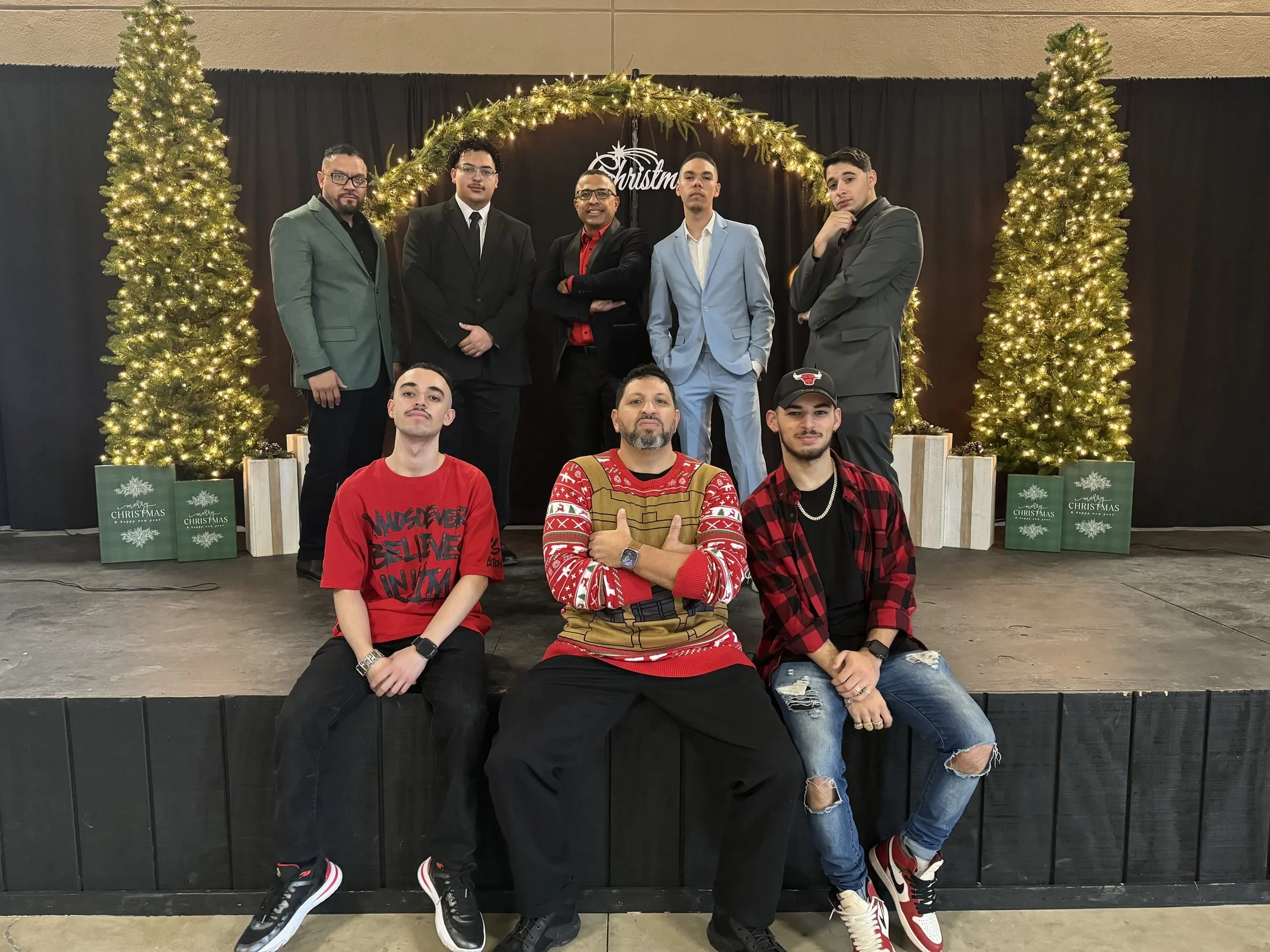 A group of seven men posing in front of a Christmas-themed backdrop with two decorated Christmas trees and a garland, some seated and some standing, wearing holiday and formal attire.