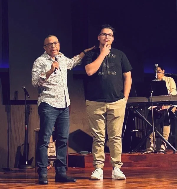 Two men standing onstage, with a musician at a keyboard and microphone in the background, engaged in a conversation or presentation.
