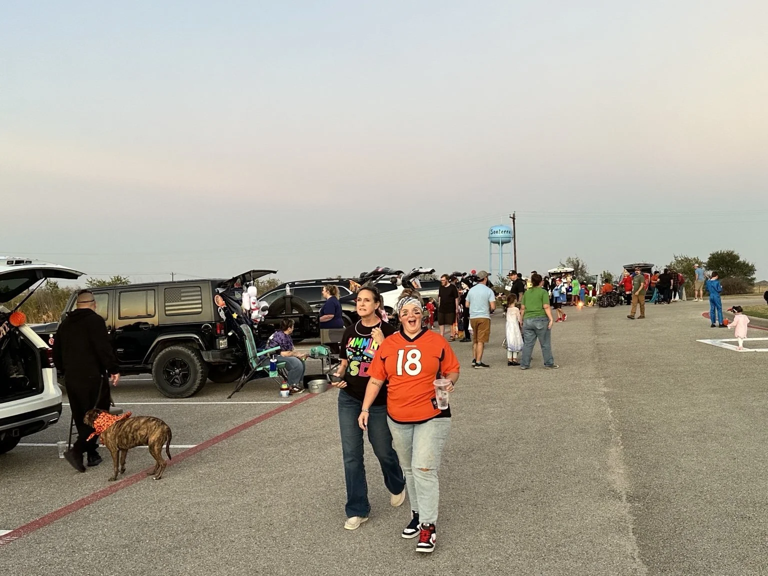 A gathering of people in an outdoor parking lot with cars, some with open trunks, and small groups socializing near makeshift tables and chairs. Two women are walking toward the camera, smiling and holding drinks, with one wearing an orange football 