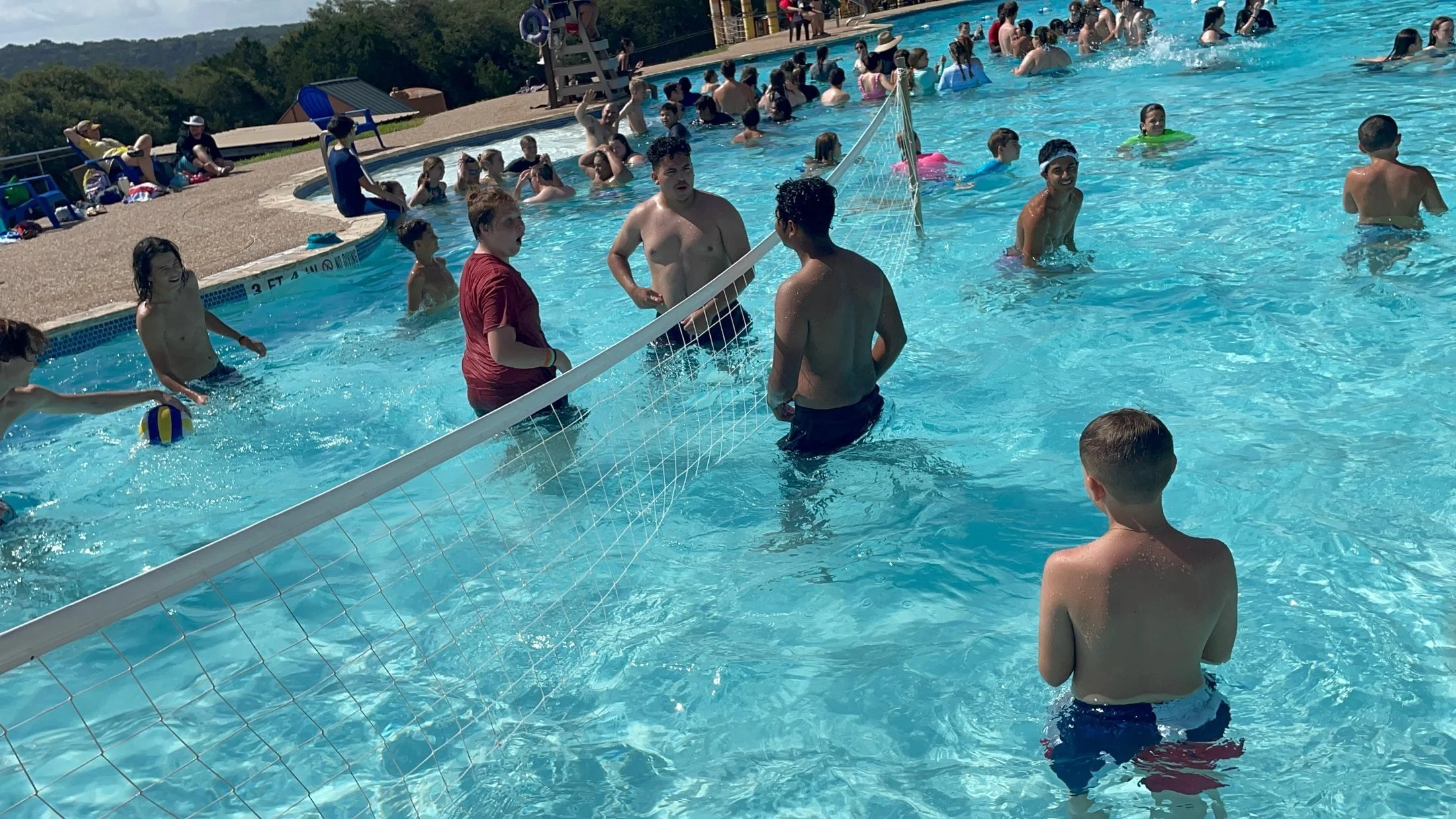 Crowded public swimming pool with children and adults swimming, playing, and relaxing on a sunny day, with hills in the background.