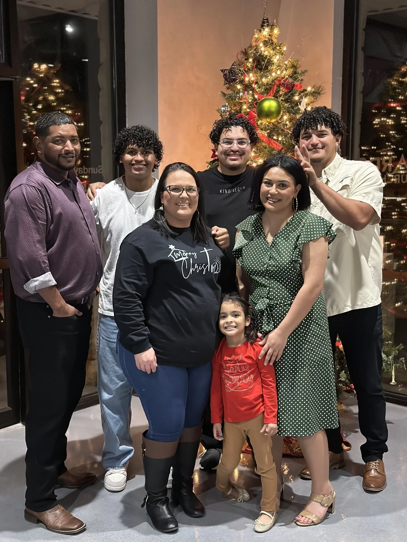 A group of seven people, including children, standing in front of a decorated Christmas tree with lights and ornaments, all smiling or posing for the photo.