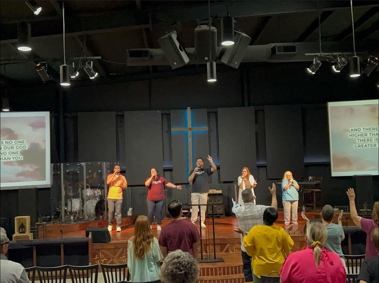 People on stage leading worship in a church with a large illuminated cross behind them. The congregation is standing and raising their hands. Two screens display song lyrics.