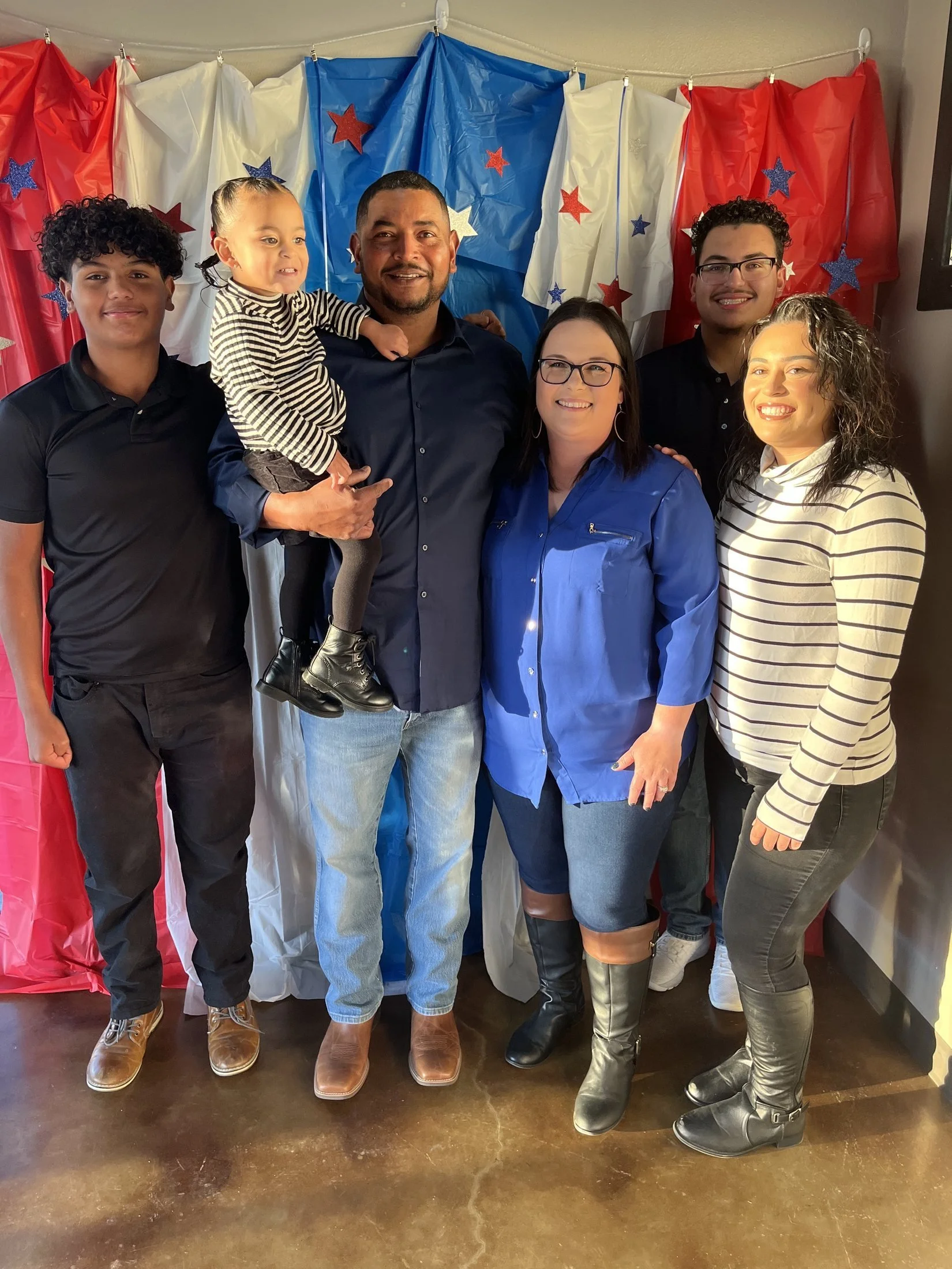 Group of six people standing in front of patriotic red, white, and blue paper backdrop with star decorations, smiling at the camera.