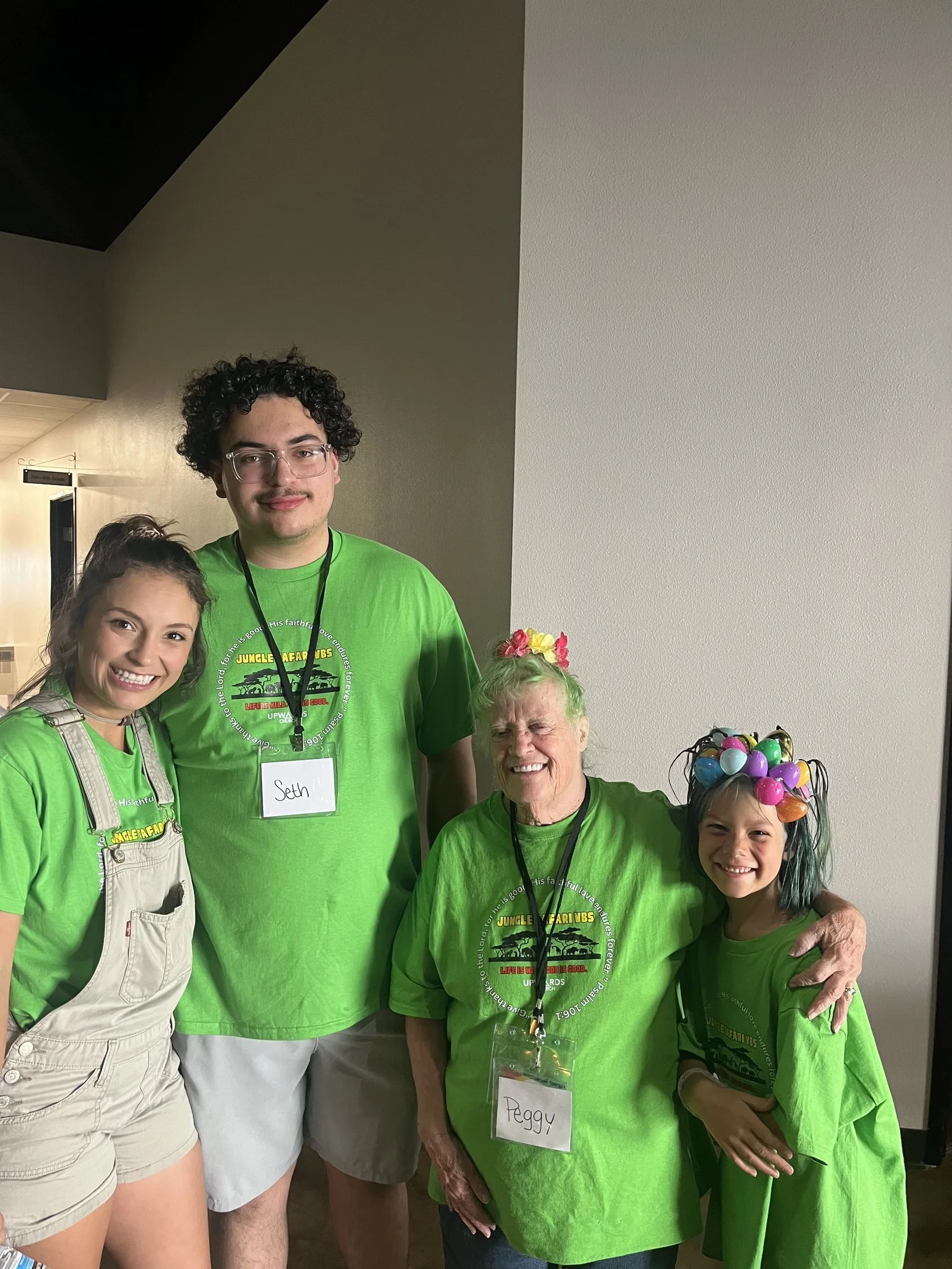 Group of five smiling people wearing green shirts, standing indoors against a plain wall, some with name tags, one person with colorful balloon headband.