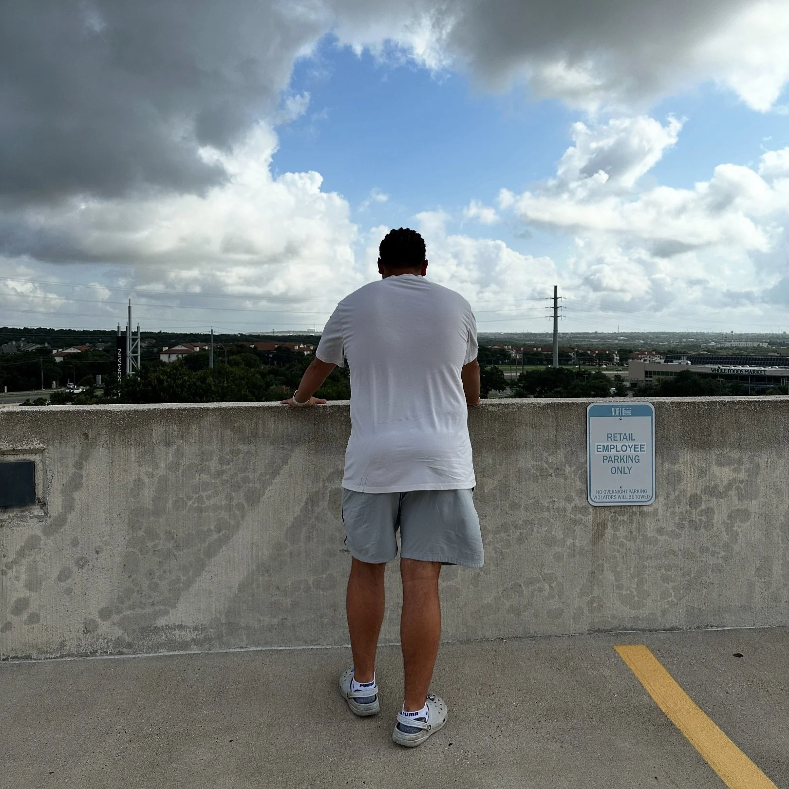 A man with short dark hair wearing a white t-shirt, gray shorts, and white sneakers standing on a rooftop parking lot, looking out over the city skyline with trees, buildings, and power lines under a partly cloudy sky.