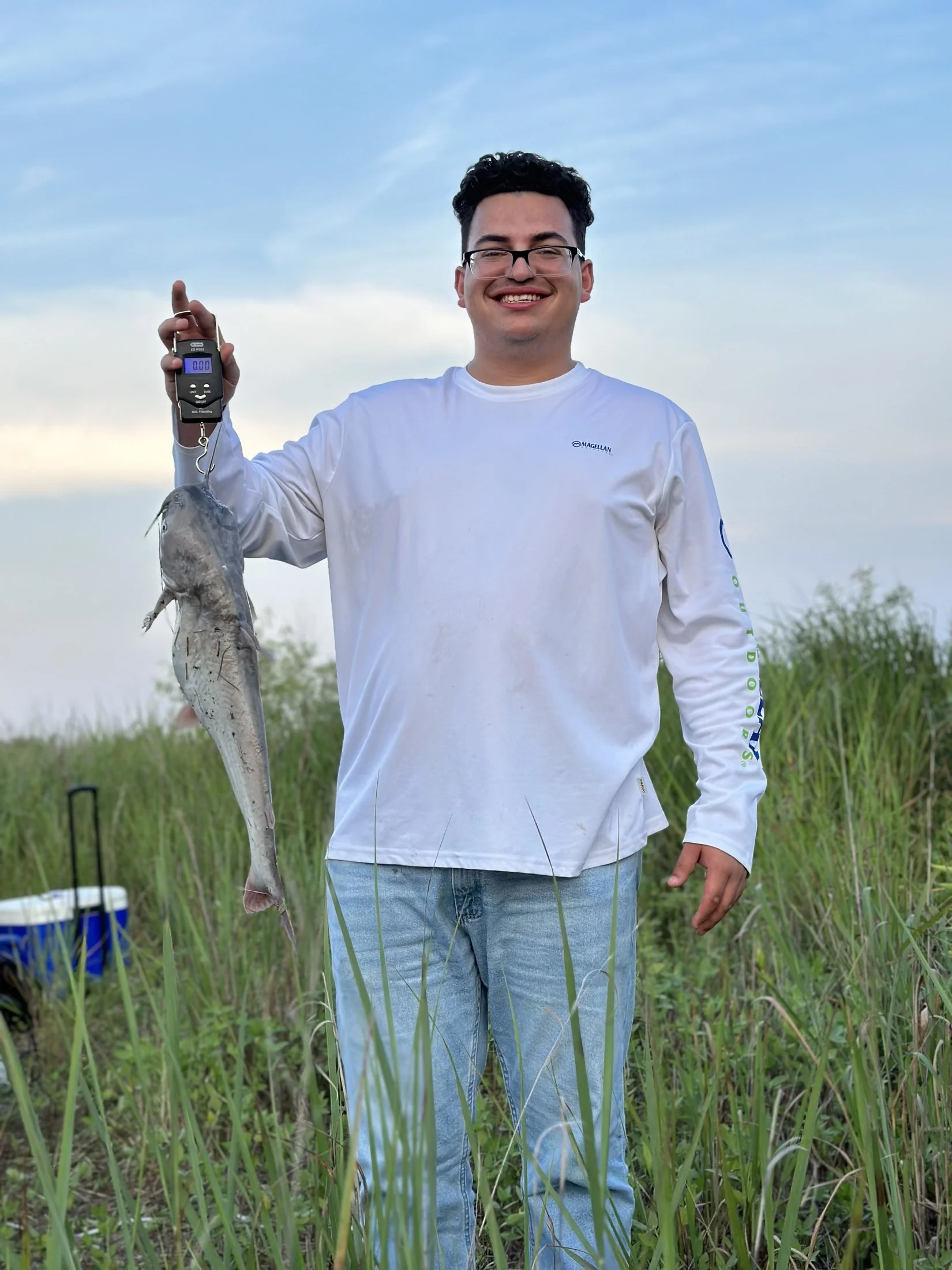 A young man with glasses and curly hair standing in a grassy field, smiling and holding a scale with a fish hanging from it.