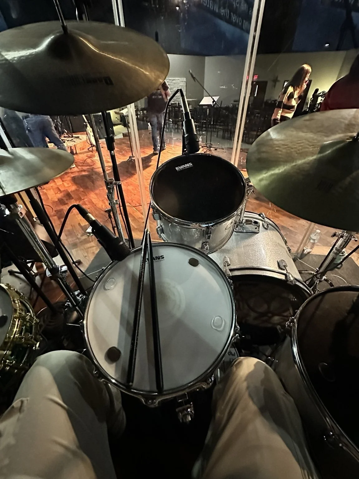 View from drummer's seat looking at drum set with microphones and cymbals, stage with musicians and a woman in the background.