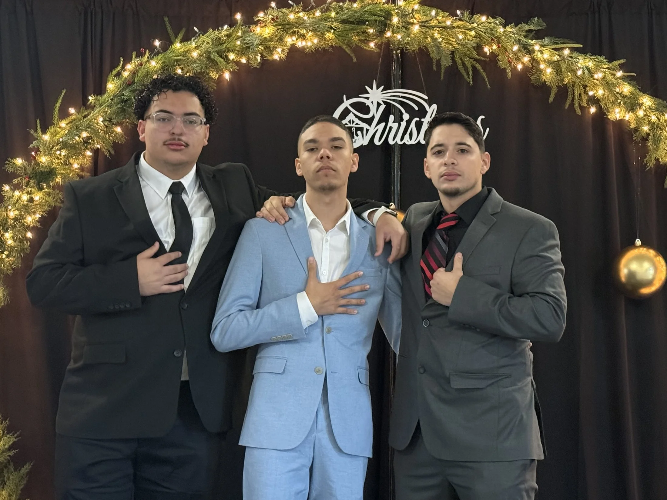 Three men in formal suits posing together at a festive event. The background features a black curtain, holiday decorations including a letter sign saying 'Christmas,' and a greenery arch with string lights.
