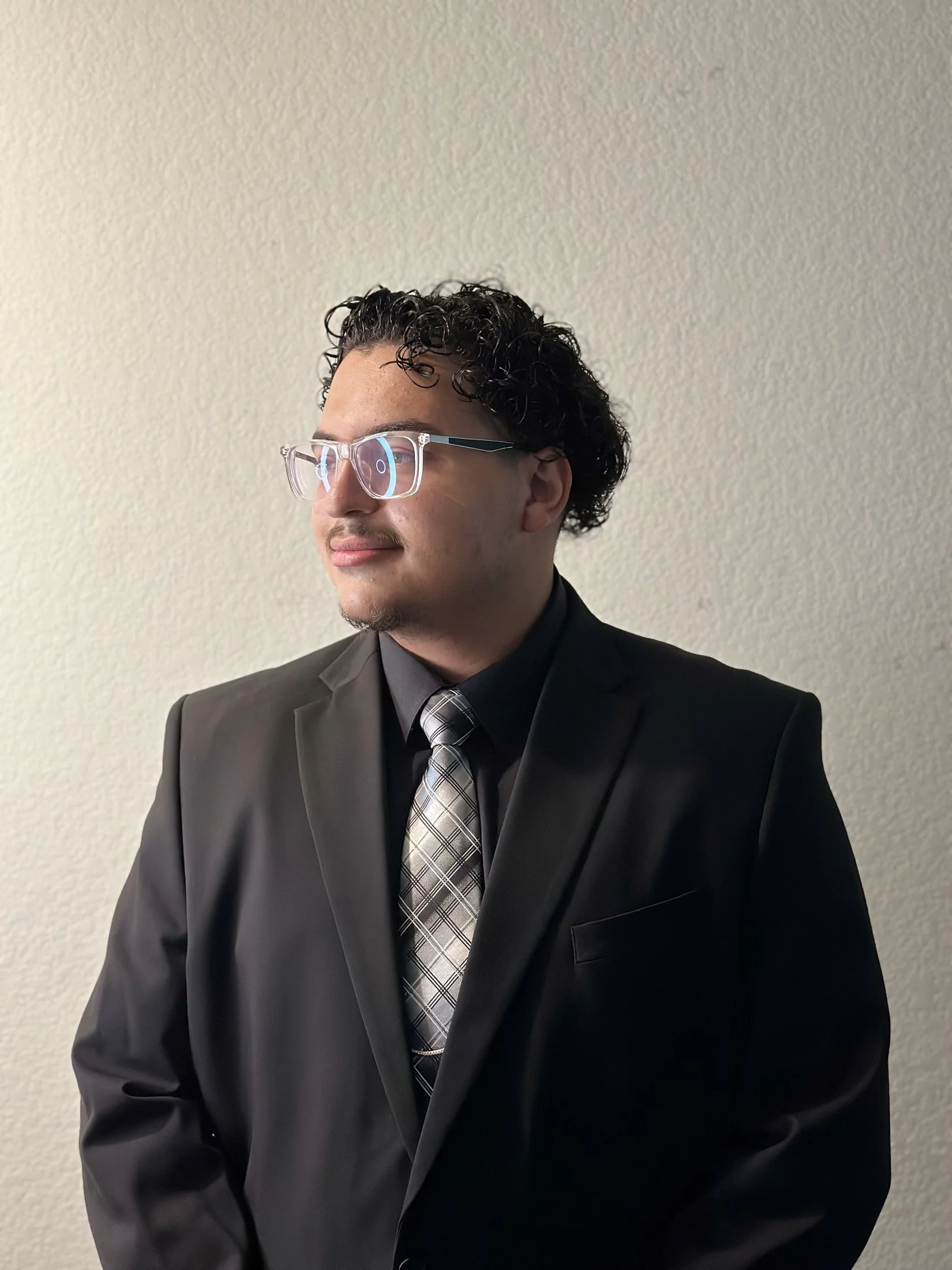 A man with curly black hair and glasses wearing a black suit and a patterned tie, standing against a plain beige wall.