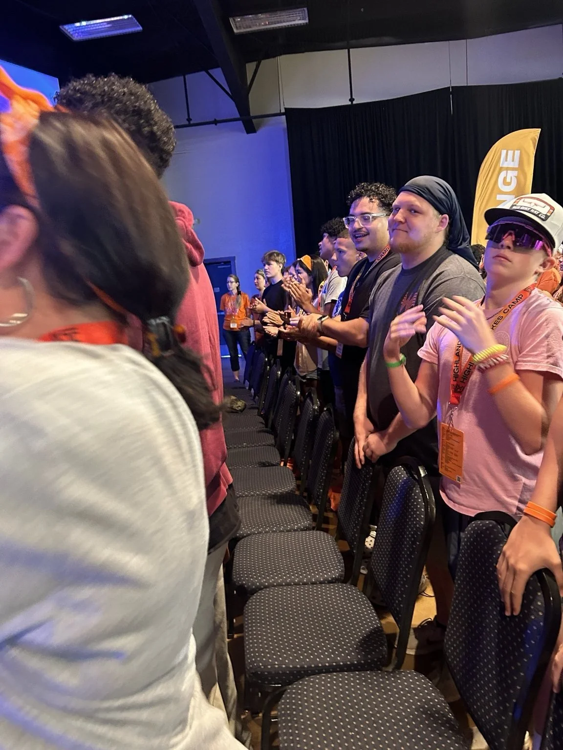 People standing in a row in an indoor auditorium, some clapping, with a black curtain and yellow banners in the background.
