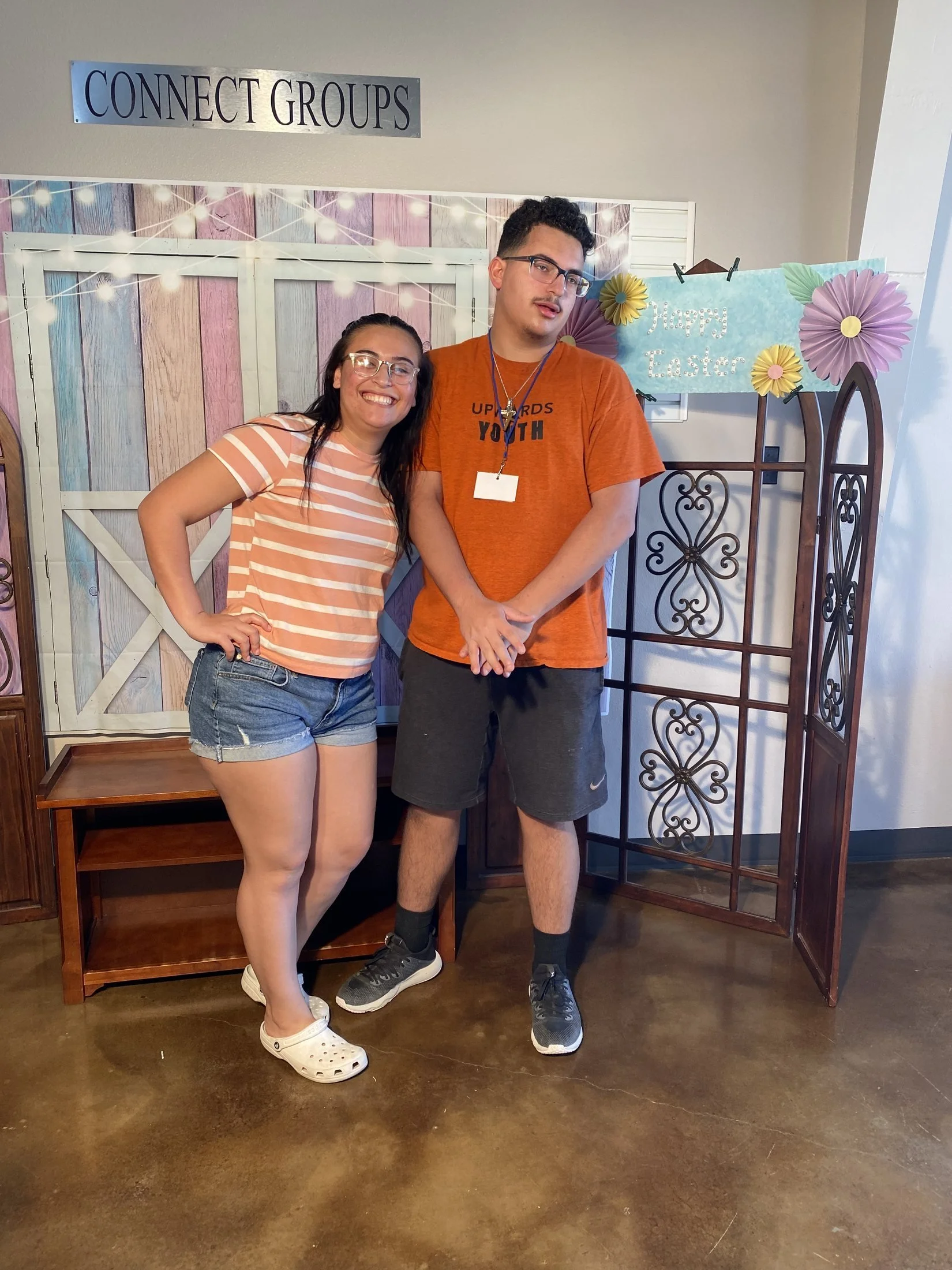 A smiling woman and a neutral man standing together in front of Easter decorations, including a blue sign that says "Happy Easter" and paper flowers.