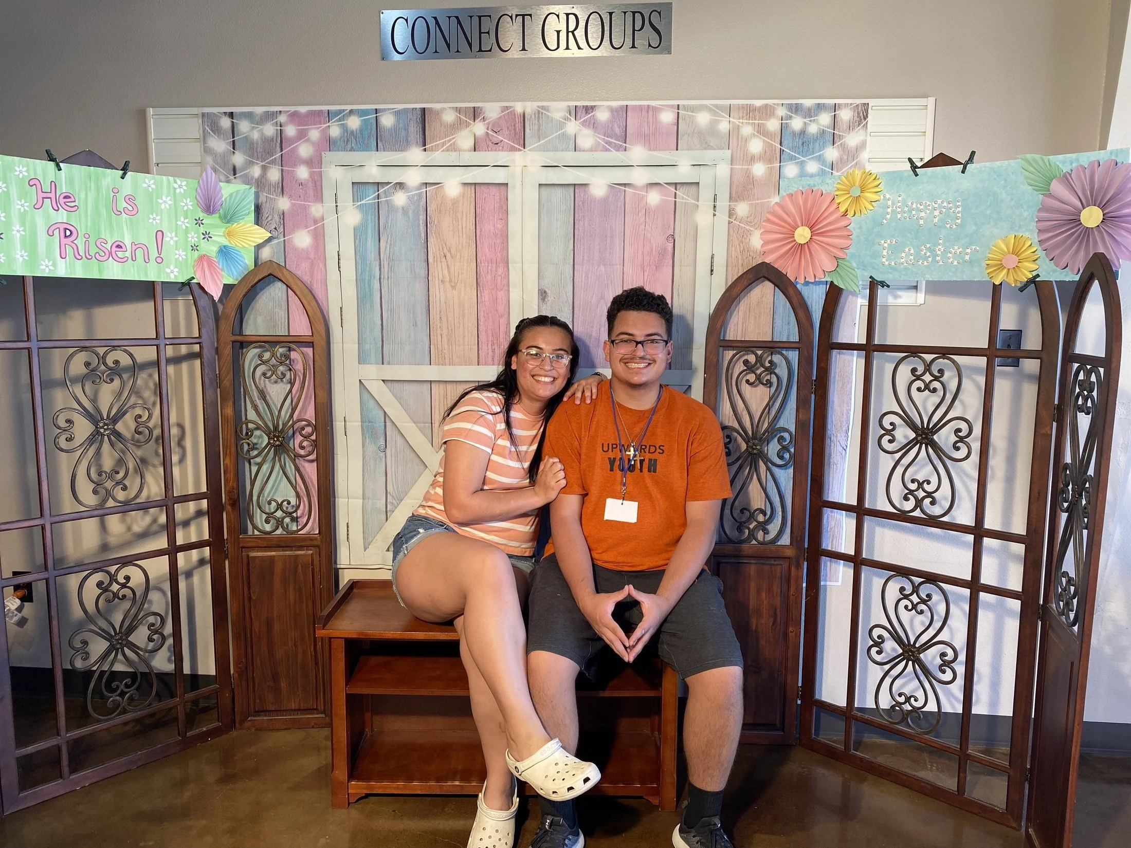 A young woman and a young man sitting close together on a wooden bench in front of a festive Easter display, smiling at the camera. The display features pastel-colored banners with Easter messages and large paper flowers, set against a backdrop of wo