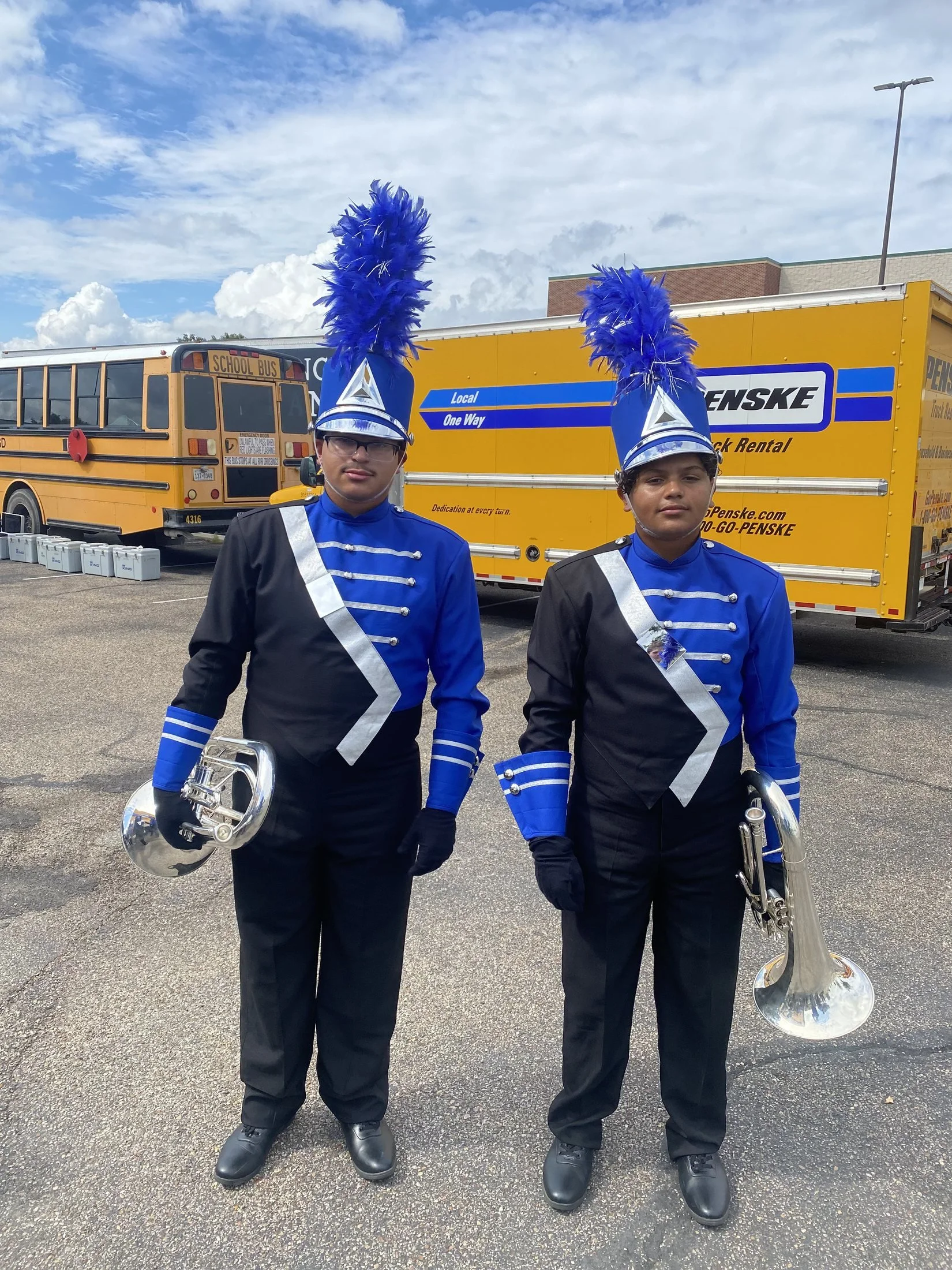 Two marching band members in uniform with tall blue feathered hats and silver accents, standing in a parking lot with a yellow school bus and Penske truck in the background.