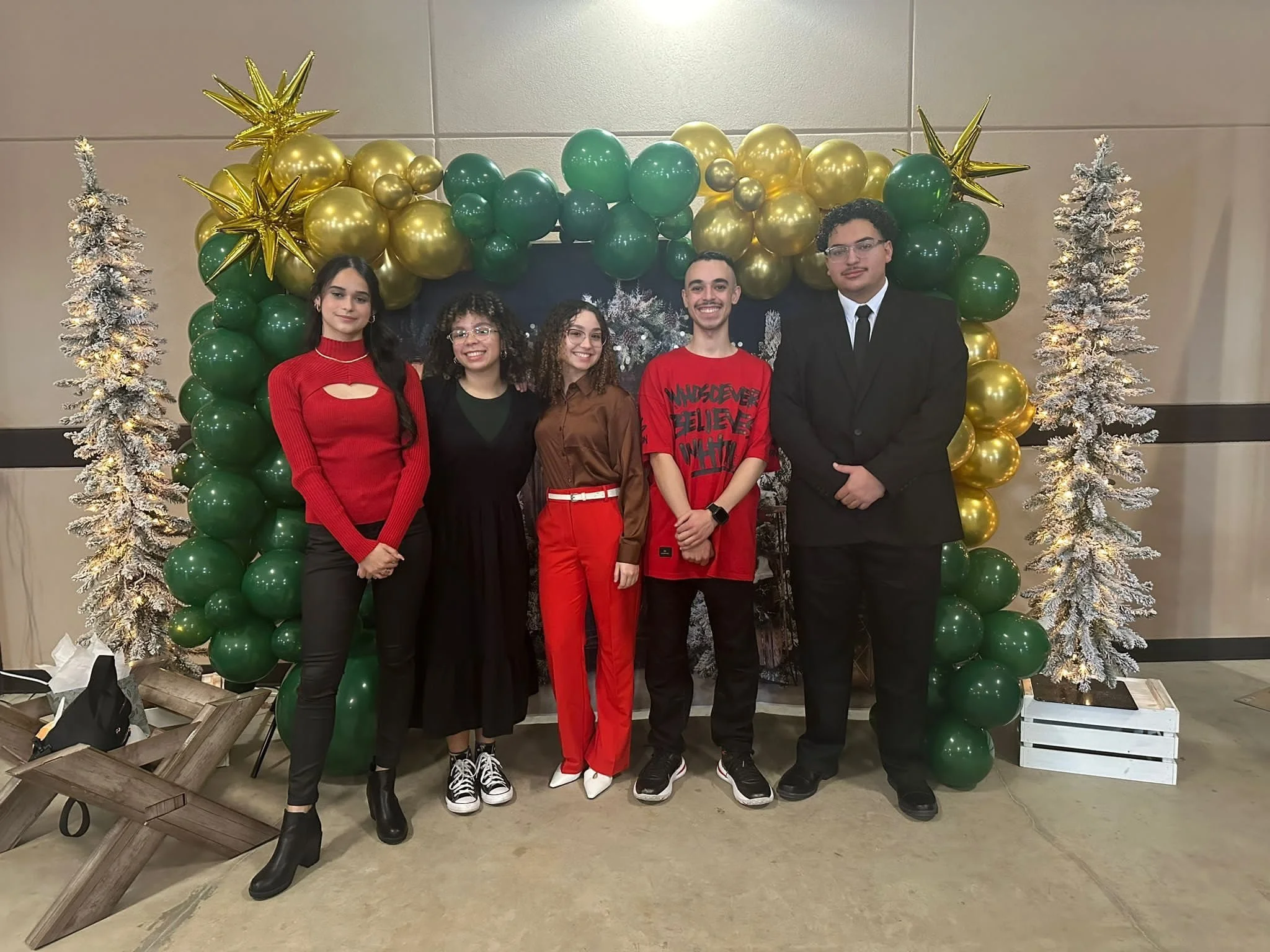 Group of five young adults standing in front of a festive holiday balloon arch with gold and green balloons, two white snow-covered Christmas trees, and holiday decor.